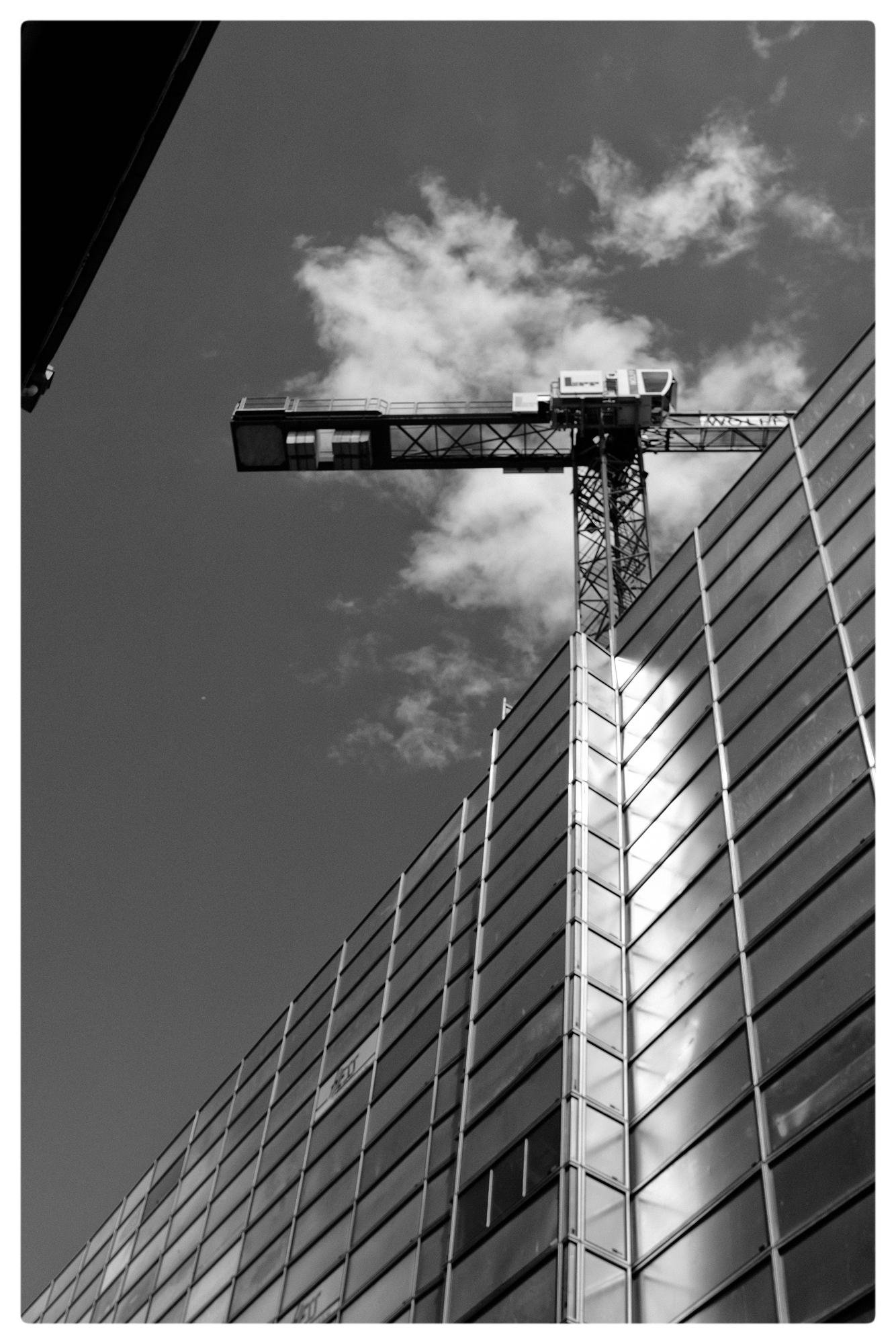 Black and white image of a glass-clad building corner with a construction crane visible in the background. The crane extends horizontally across the sky, with scattered clouds providing contrast against the darker sky. Reflections of light are visible on the building's windows.