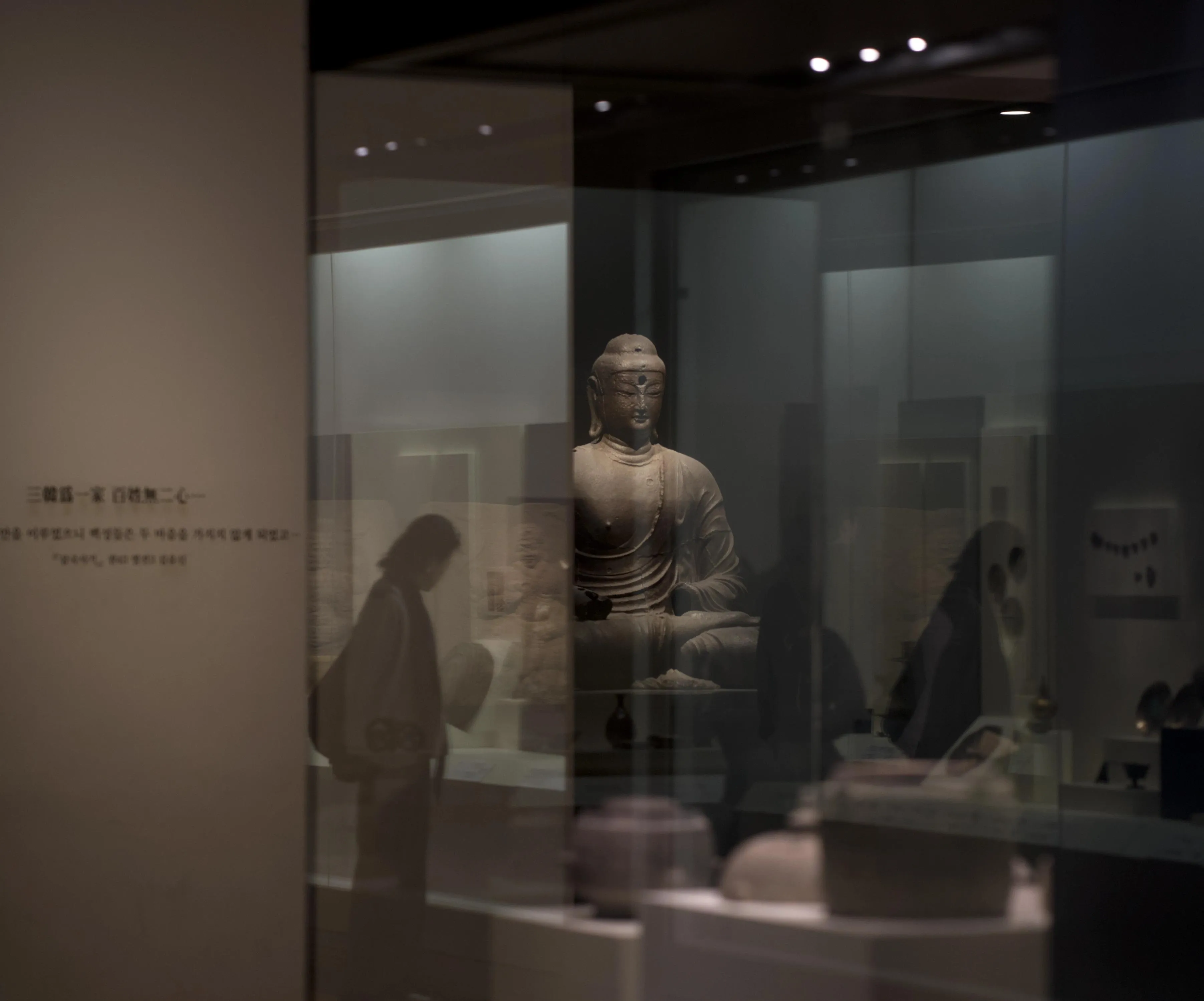Photograph of a stone statue of Buddha exhibited in a museum. The statue is seen through a glass surface reflecting other museum artifacts such as ceramic pots, metallic wares and stone reliefs, as well as the silhouettes of museum visitors looking at these artifacts.