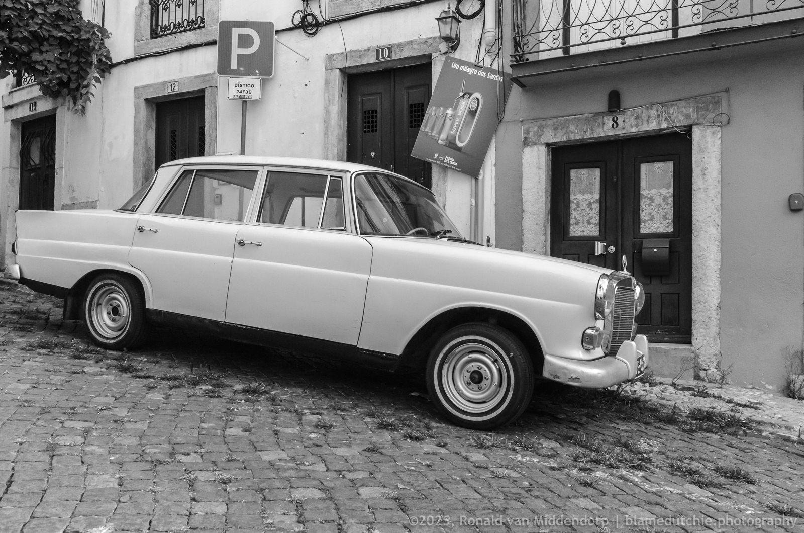 Black-and-white photo of a vintage four-door Mercedes parked on a cobblestone street beside old building facades with numbered doorways, a “P” parking sign, and a hanging shop sign.