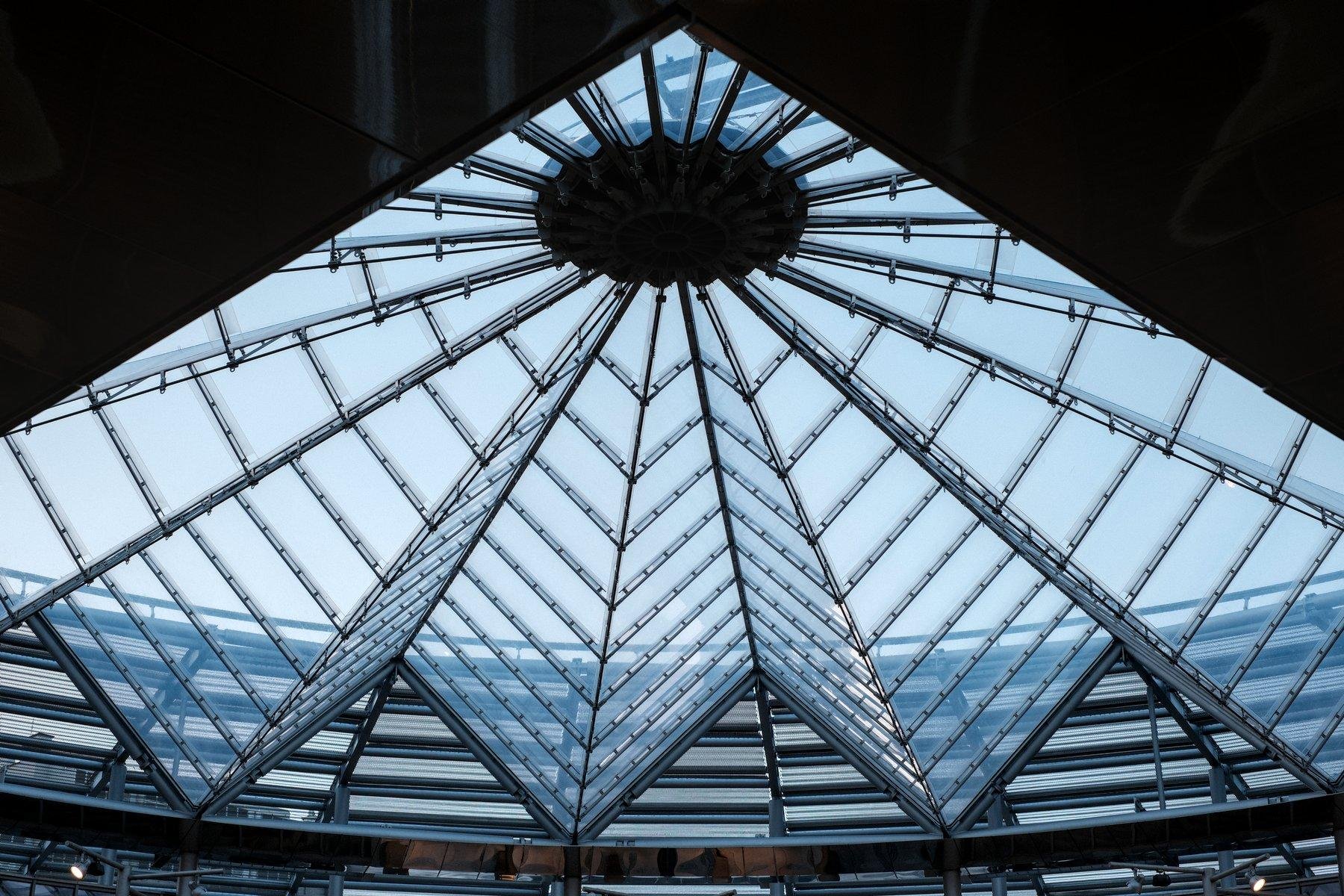 Photo horizontale. Contre plongée sur le toit en verre de la station Villejuif Gustave Roussy. En haut, en contre jour, des bouts d’escalator formant un masque sur la verrière, qui avec sa forme fait penser à un éventail de verre