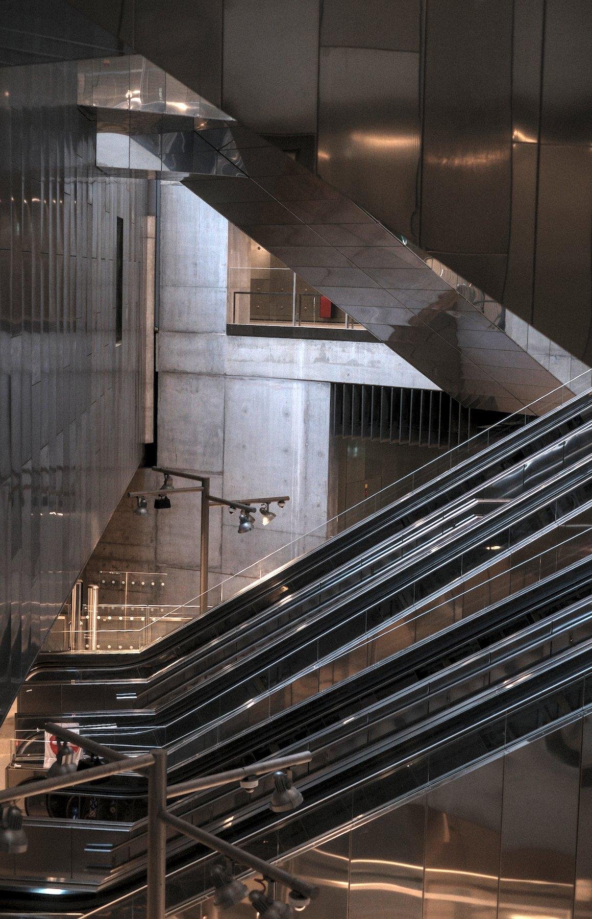 Photo verticale. En bas, des escalators vu depuis un étage au dessus, un mur en métal à gauche qui laisse place à un plafond au niveau de ces escalators, et en haut la paroi latérale d’escalators de l’étage au dessus, les 3 éléments délimitant un triangle dans l’image