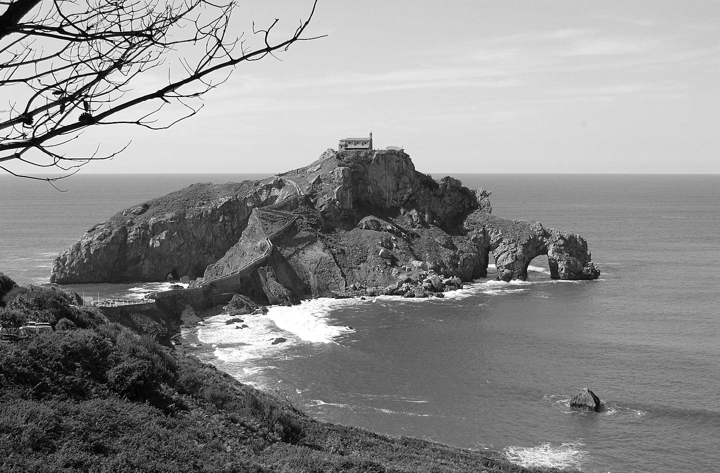 Una isla rocosa con una ermita en su punto más alto y un puentecito que da acceso a ella.
Está cerca de la costa.
En el primer plano, arriba a la izquierda unas ramas de un árbol enmarcan la imagen.
Tras ella el Cantábrico.