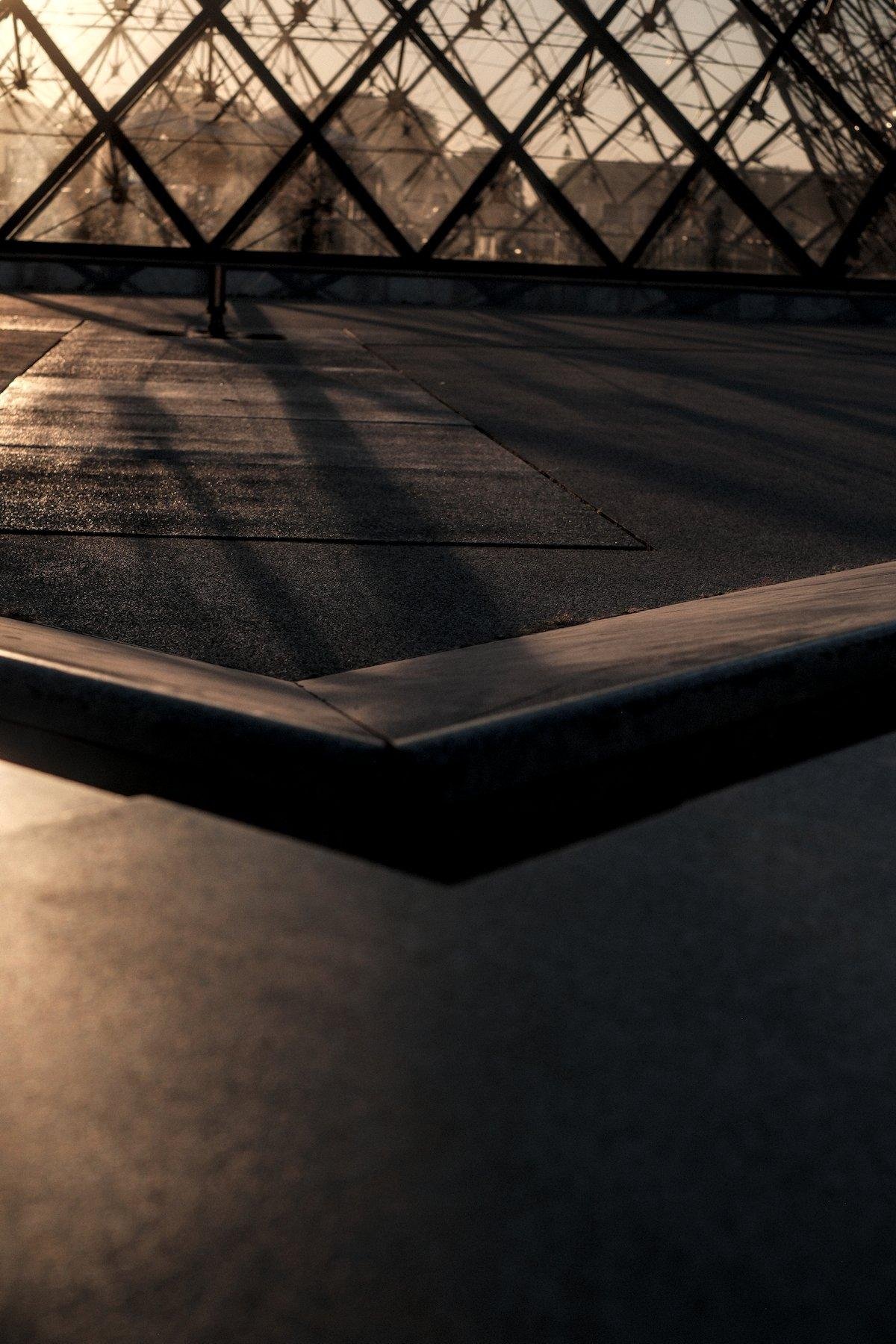 Photo verticale. Le sol puis au dessus le coin d’une fontaine du Louvre et en haut, le motif caractéristique d’une des paroie de la pyramide du Louvre, éclairés par un soleil couchant venant d’en haut à gauche