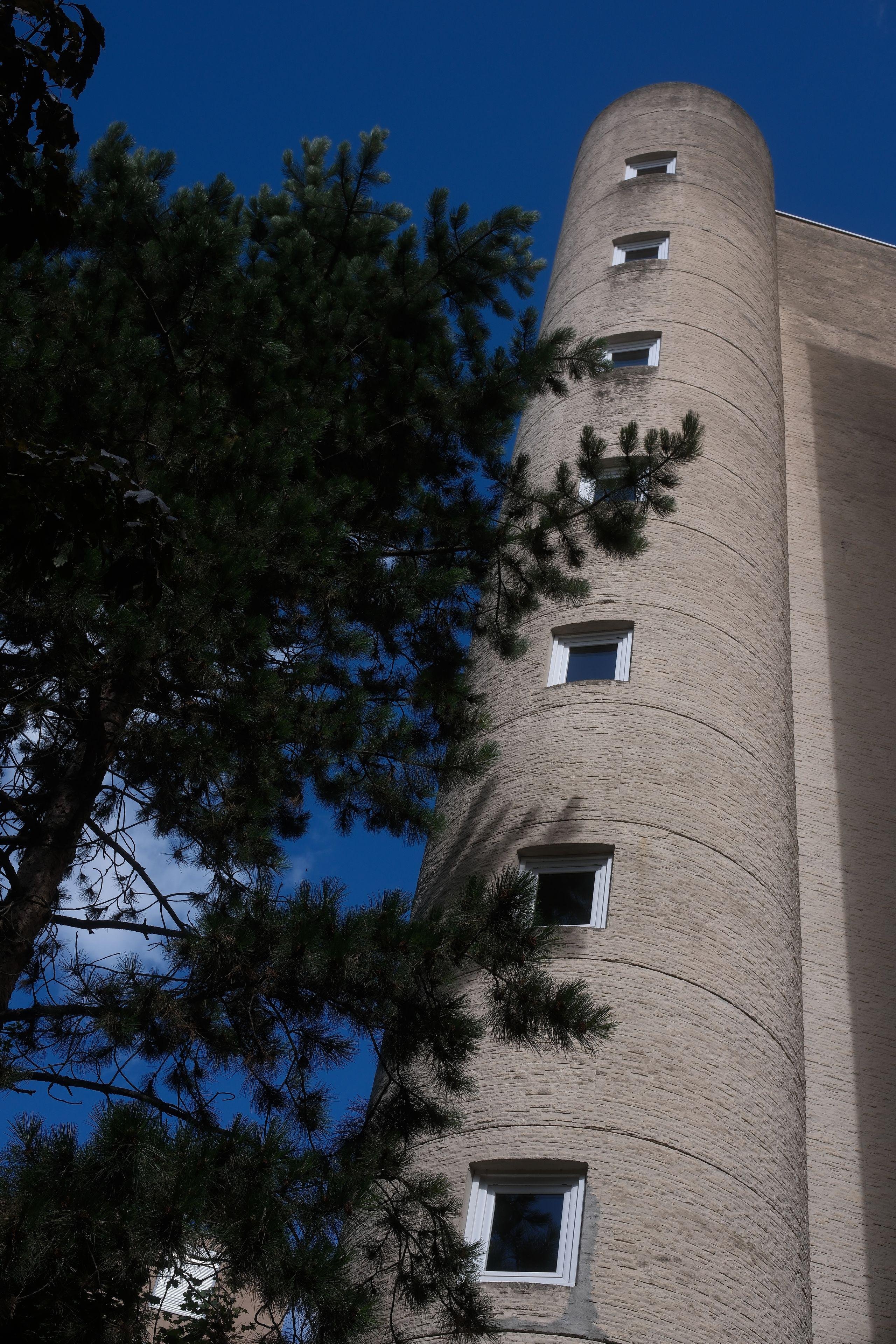 A tower of a building, probably staircases, with regular vertical windows, from a low angle, towards the deep blue sky. Some trees on the left side of the pic
