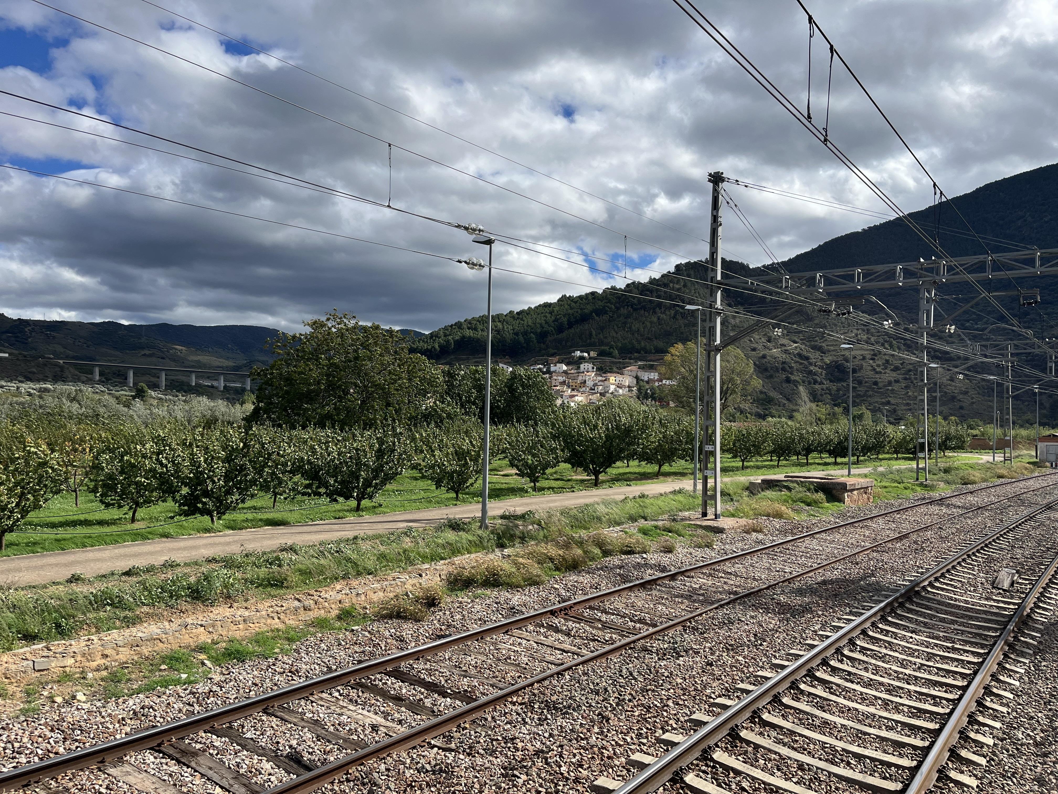 The image shows a railway track with overhead electric lines in a rural setting. There are trees, possibly an orchard, along a path next to the tracks. In the background, there are hills or mountains, and some buildings nestled among the greenery. The sky is partly cloudy.