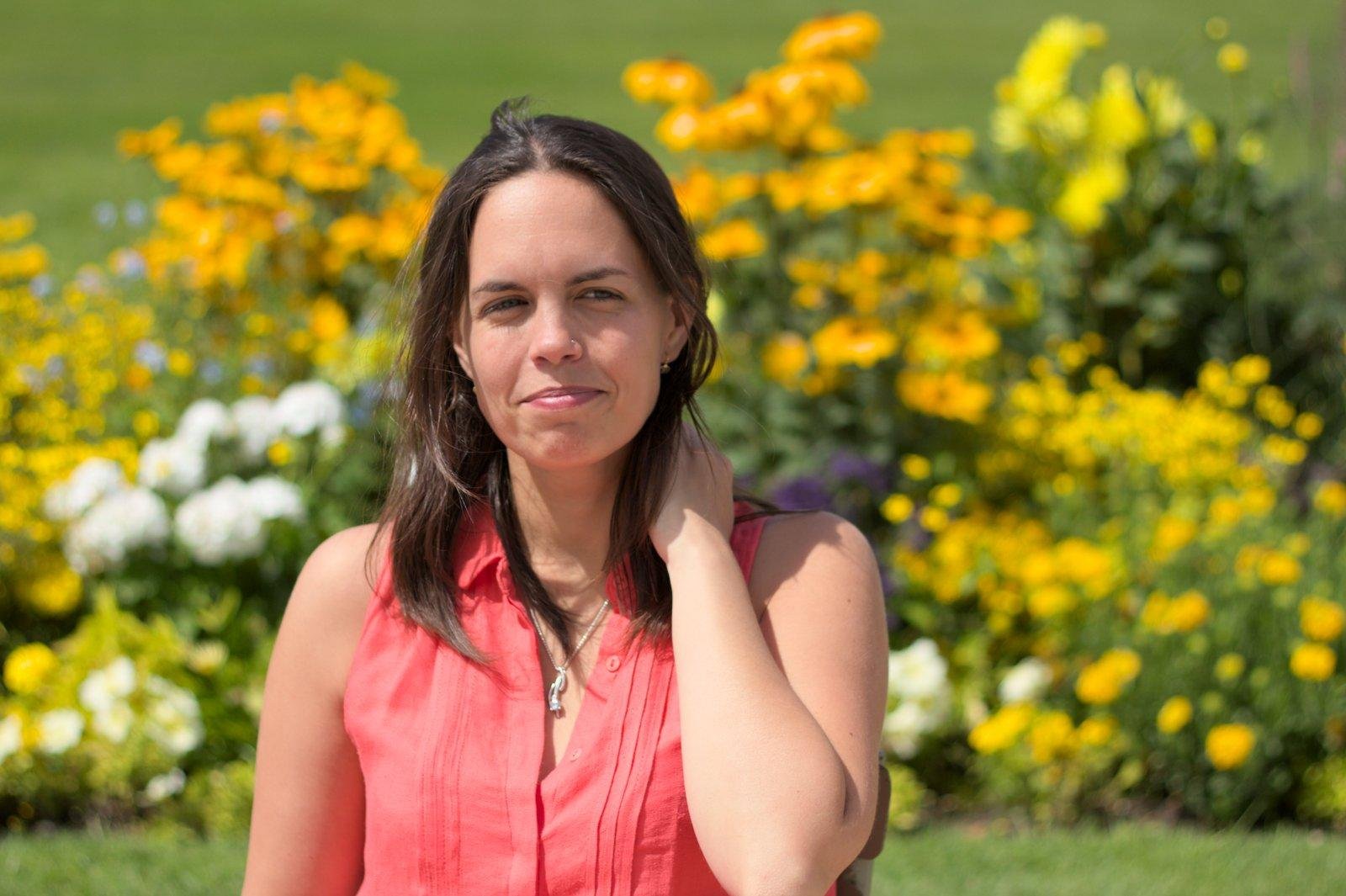 Photo format paysage montrant de face, une jeune femme brune aux cheveux long lâchés, habillée de rose/rouge sans manche avec un collier autour du coup, se tenant les cheveux contre la nuque de la main gauche, les yeux regardant vers la gauche avec un sourire un peu crispé mais joyeux. Le fond est plein de fleurs jaunes, oranges et blanches et de verdure