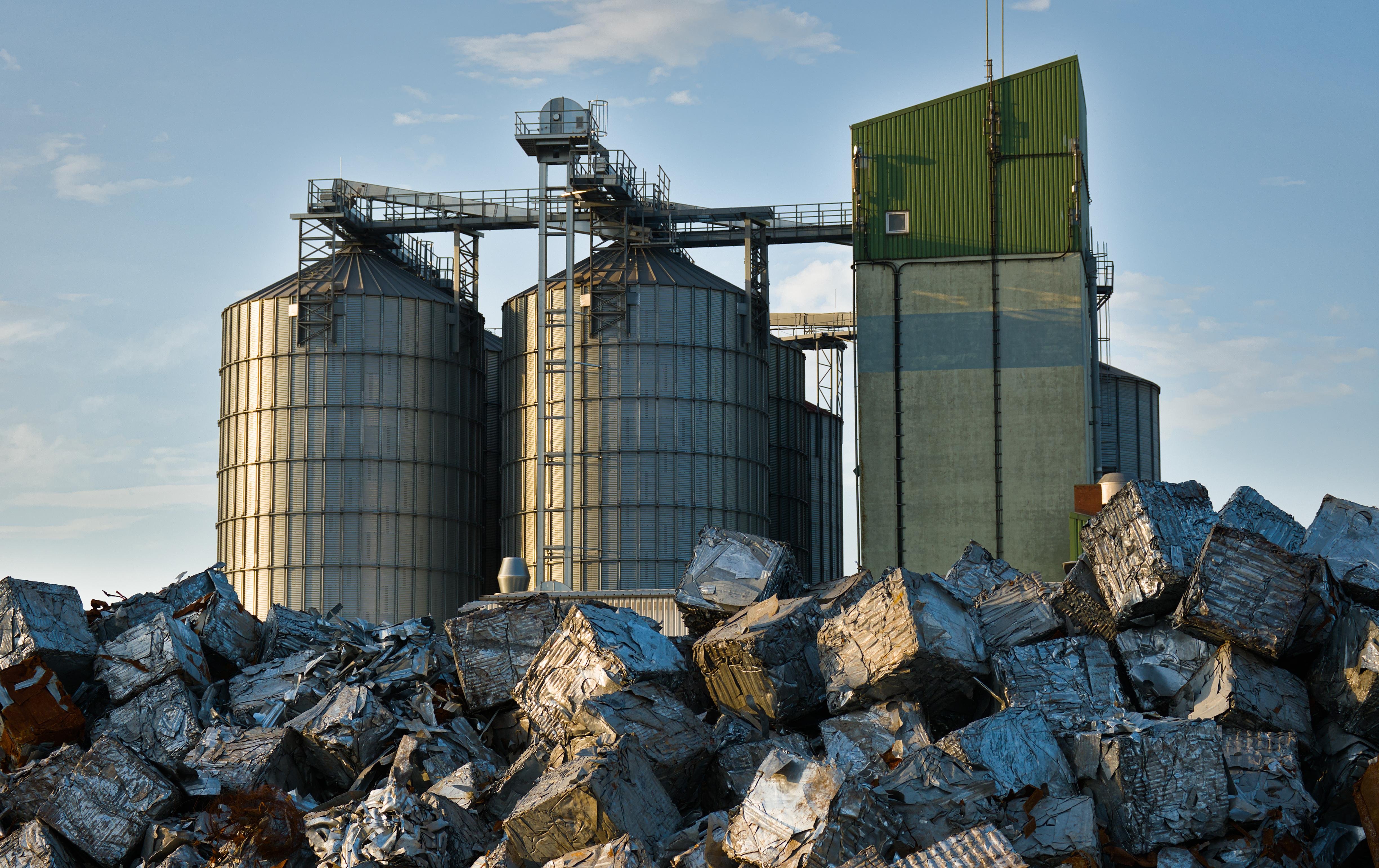 Three large industrial silos made of metal are positioned side by side against a clear sky. A network of walkways and pipes is visible on top of the silos. In the foreground, a heap of compressed metal cubes is piled up, reflecting light from the sun. A green structure with vertical stripes and a small antenna is situated to the right of the silos. 
### 
Drei große Industriesilos aus Metall sind Seite an Seite gegen einen klaren Himmel positioniert. Ein Netz von Gehwegen und Rohren ist auf den Silos zu sehen. Im Vordergrund türmt sich ein Haufen aus komprimierten Metallwürfeln auf, der Licht von der Sonne reflektiert. Eine grüne Struktur mit vertikalen Streifen und einer kleinen Antenne befindet sich rechts von den Silos.