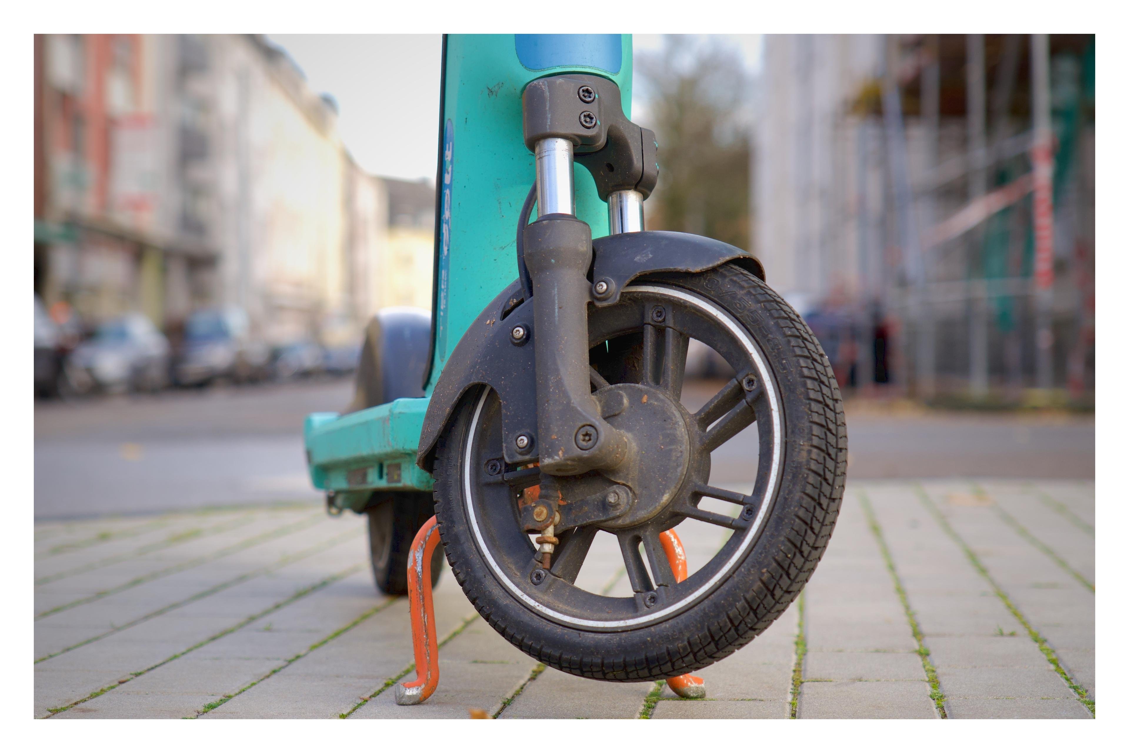 Nahaufnahme eines abgenutzten E-Scooters mit Stützständer auf grauen Pflastersteinen in unscharfer Straßenkulisse.