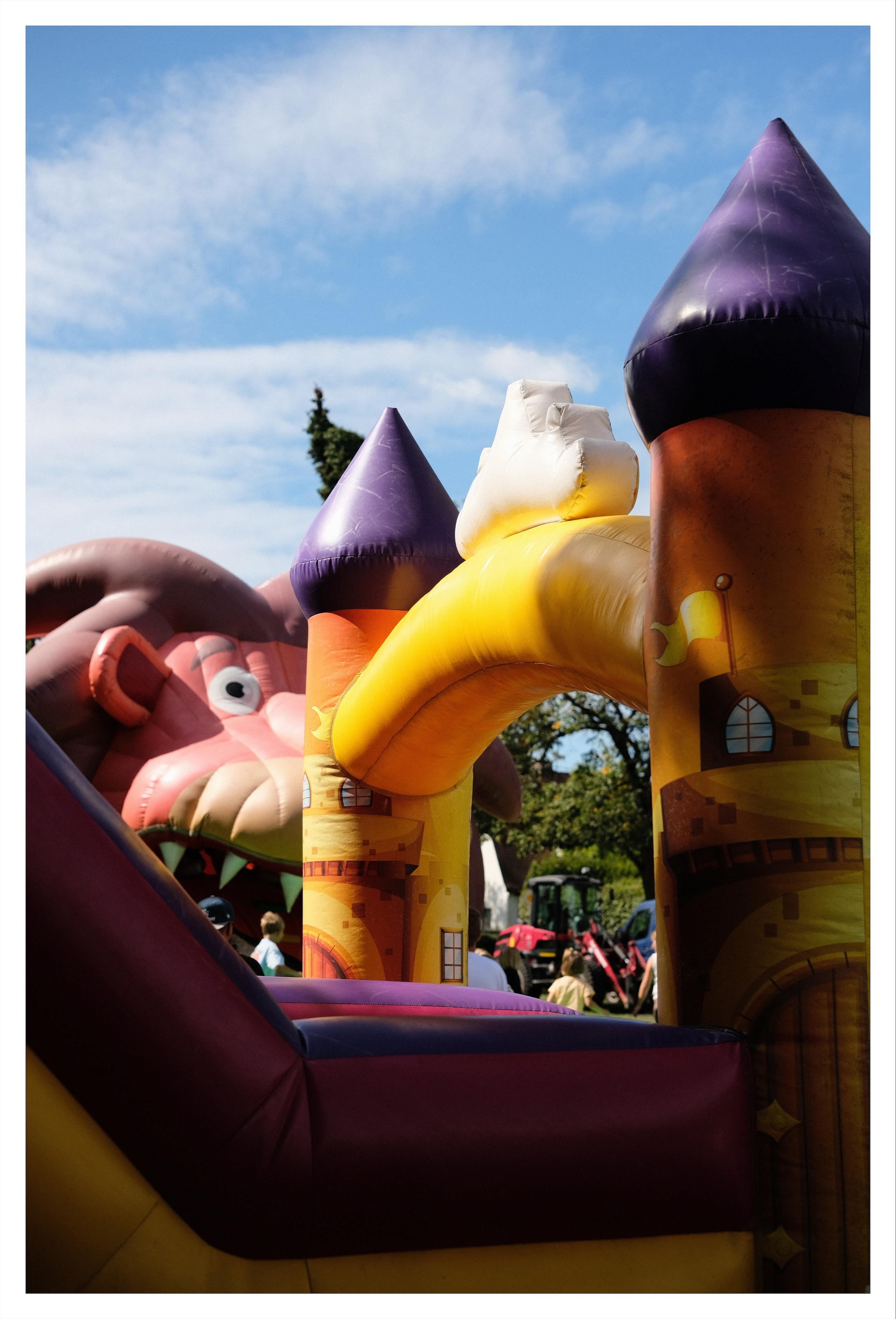 Inflatable bouncy castle with yellow arch and tall towers topped by purple and white cones, set outdoors under a blue sky; a huge inflatable animal head (likely a colourful lion) is visible in the background.