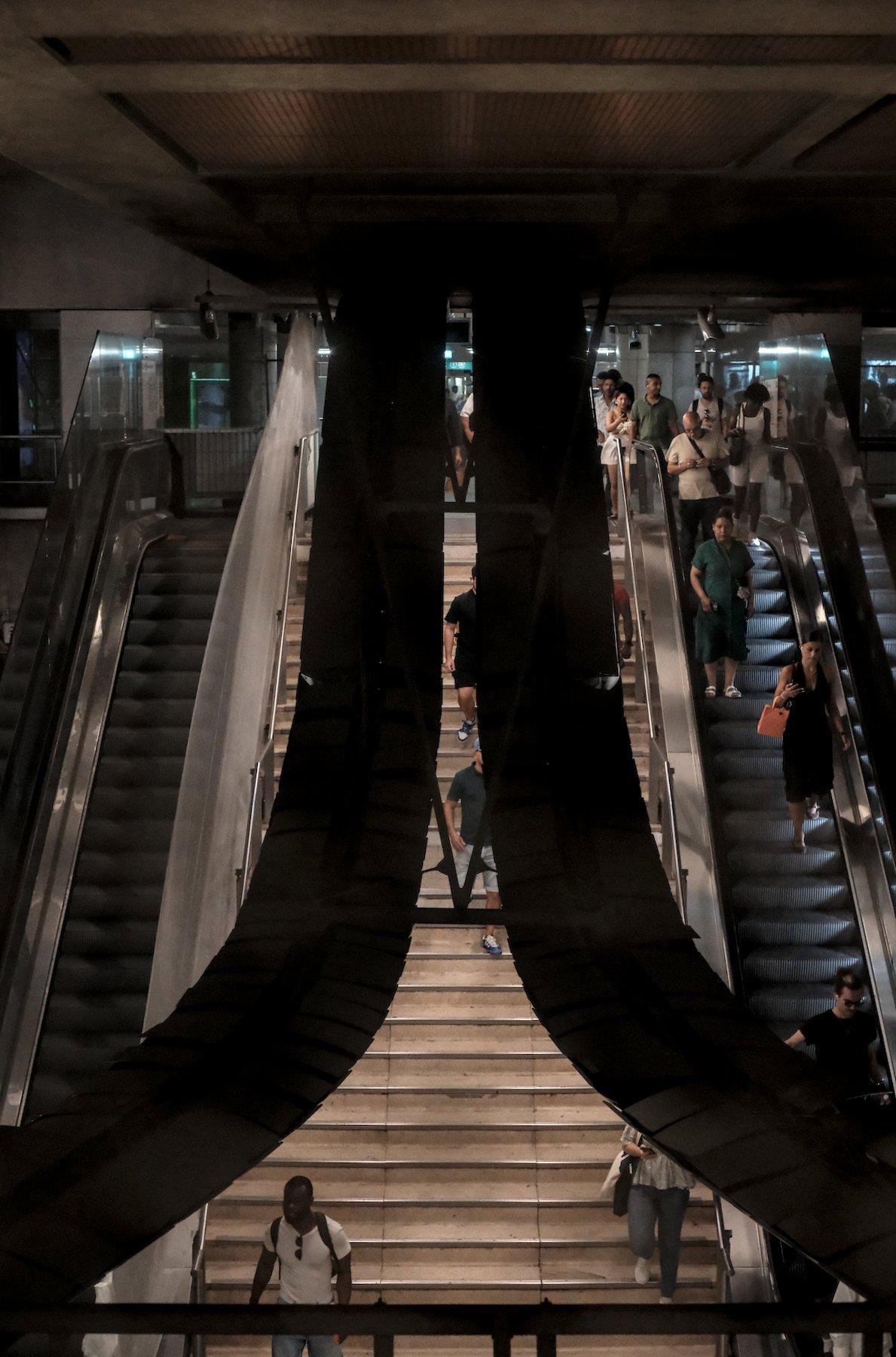 Photo verticale. À gauche, une escalator vide, en maintenance, à droite un escalator montant, avec quelques personnes dessus. Au centre, arrière plan, un escalier entre les 2 escalators. Au premier plan, la silhouette de 2 bandes arrivant d’en bas à gauche et à droite, se rejoignant au centre en courbe et qui suivent ensuite les escaliers. C’est le support des lampes d’éclairage