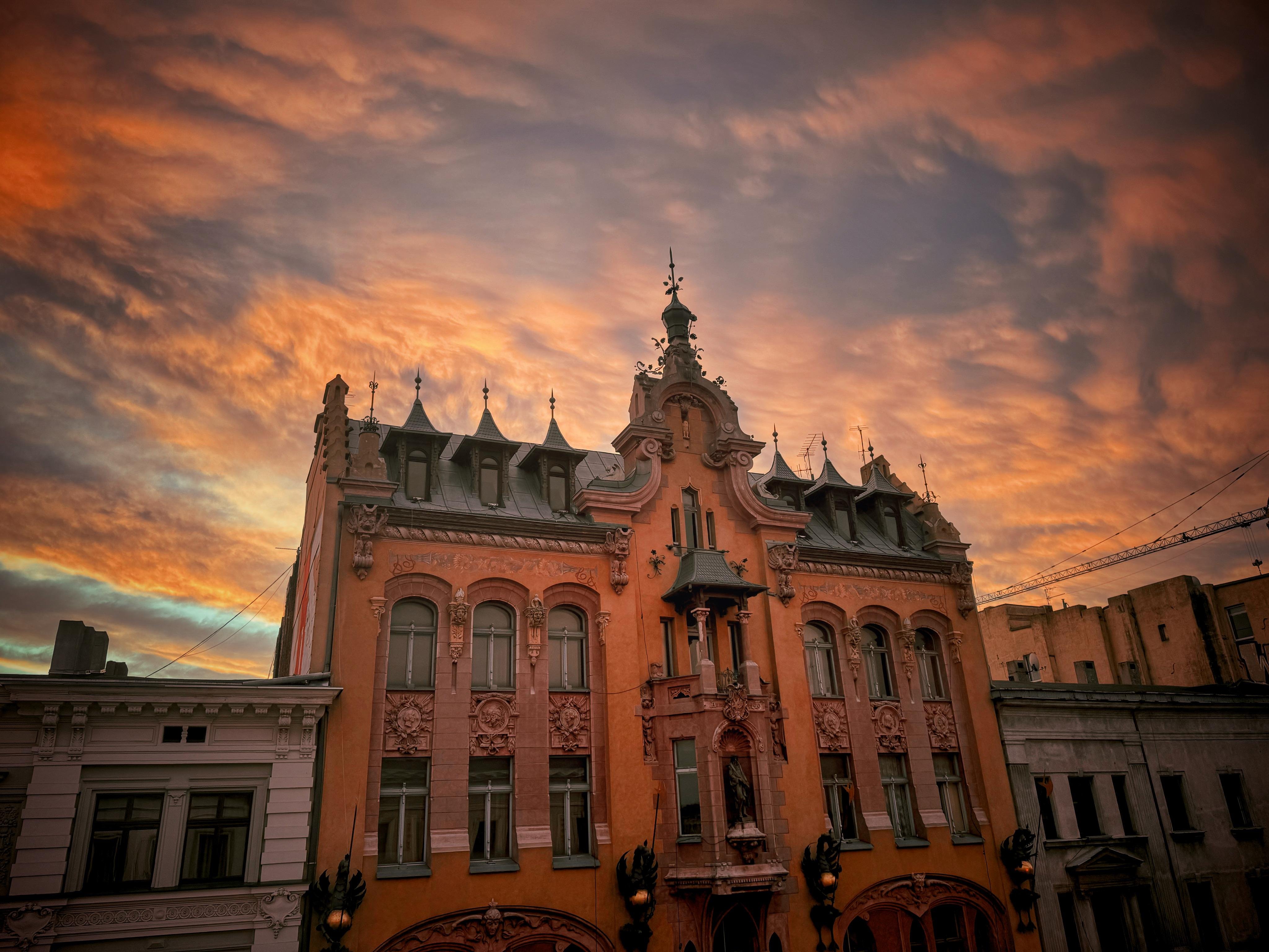 The image depicts a historic building (Tenement house under Gutenber in Łódź, Poland) with intricate architectural details against a dramatic, colorful sky at sunset. The building features multiple peaked roofs, arched windows, decorative elements, and ornate facades. The sky is filled with warm hues, including shades of orange, pink, and purple, which enhance the building's appearance.