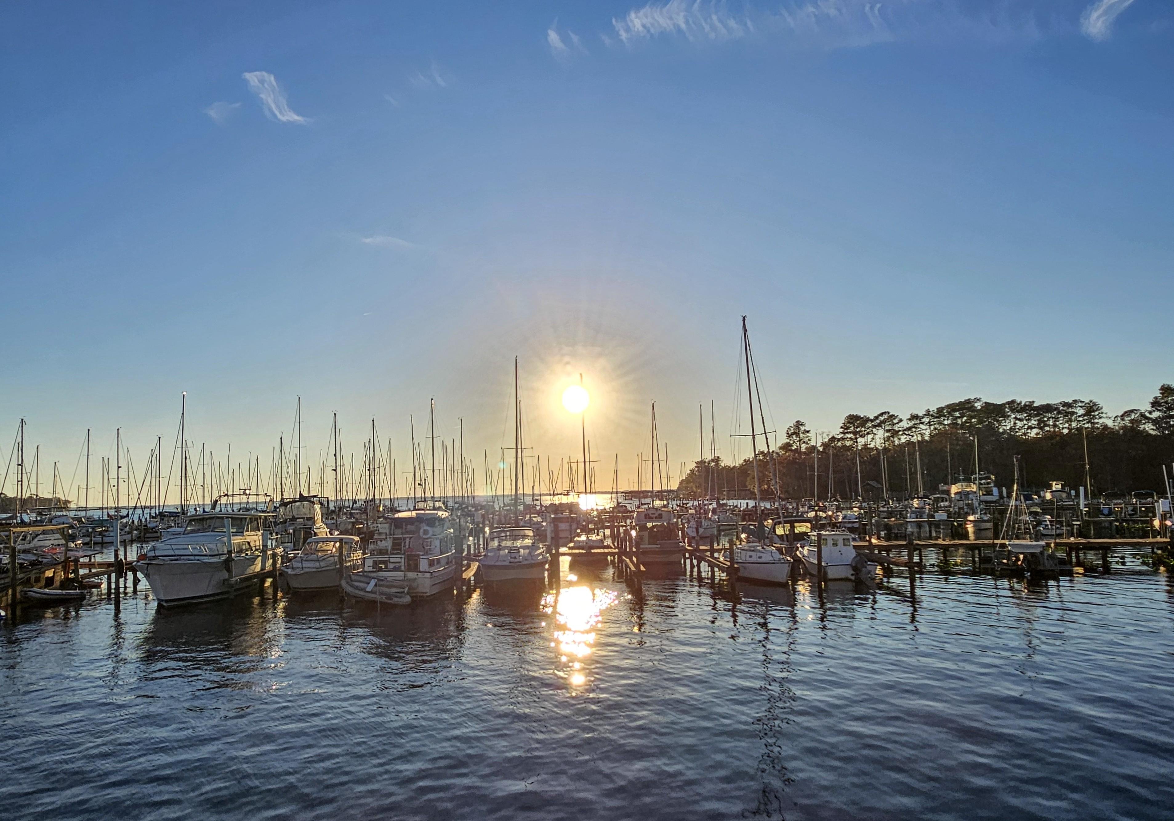 Sunset over a marina with numerous boats docked, their masts silhouetted against the sky. The sun is low on the horizon, casting a reflection on the calm water. Trees line the background on the right, with a few wispy clouds visible in the clear blue sky.