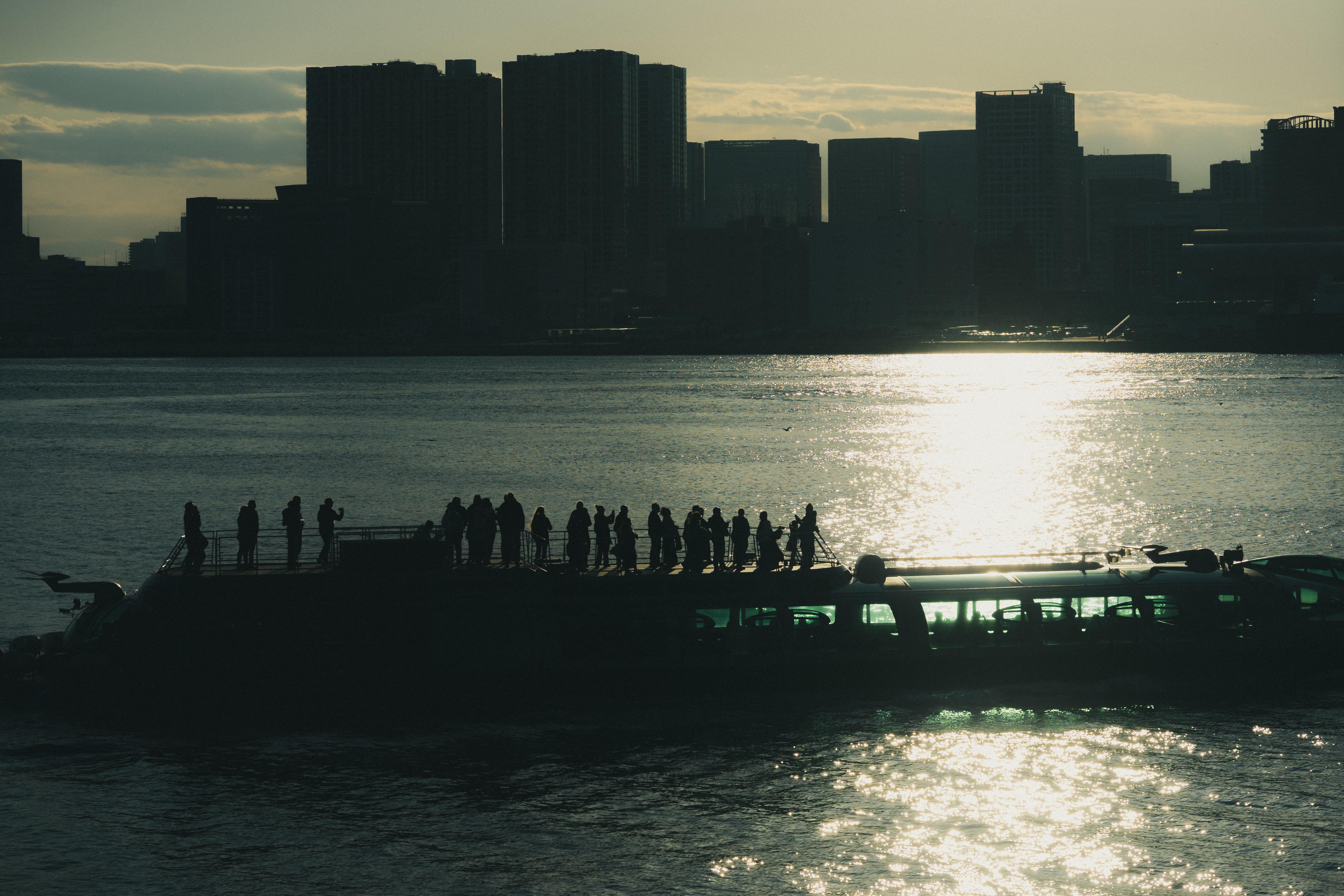 The image shows a silhouette of a boat with people standing on it, set against a city skyline. The scene is captured during sunset or sunrise, with the sun reflecting off the water, creating a dramatic and atmospheric effect. The city buildings are darkened in the background, providing a clear contrast to the brighter reflections on the water.