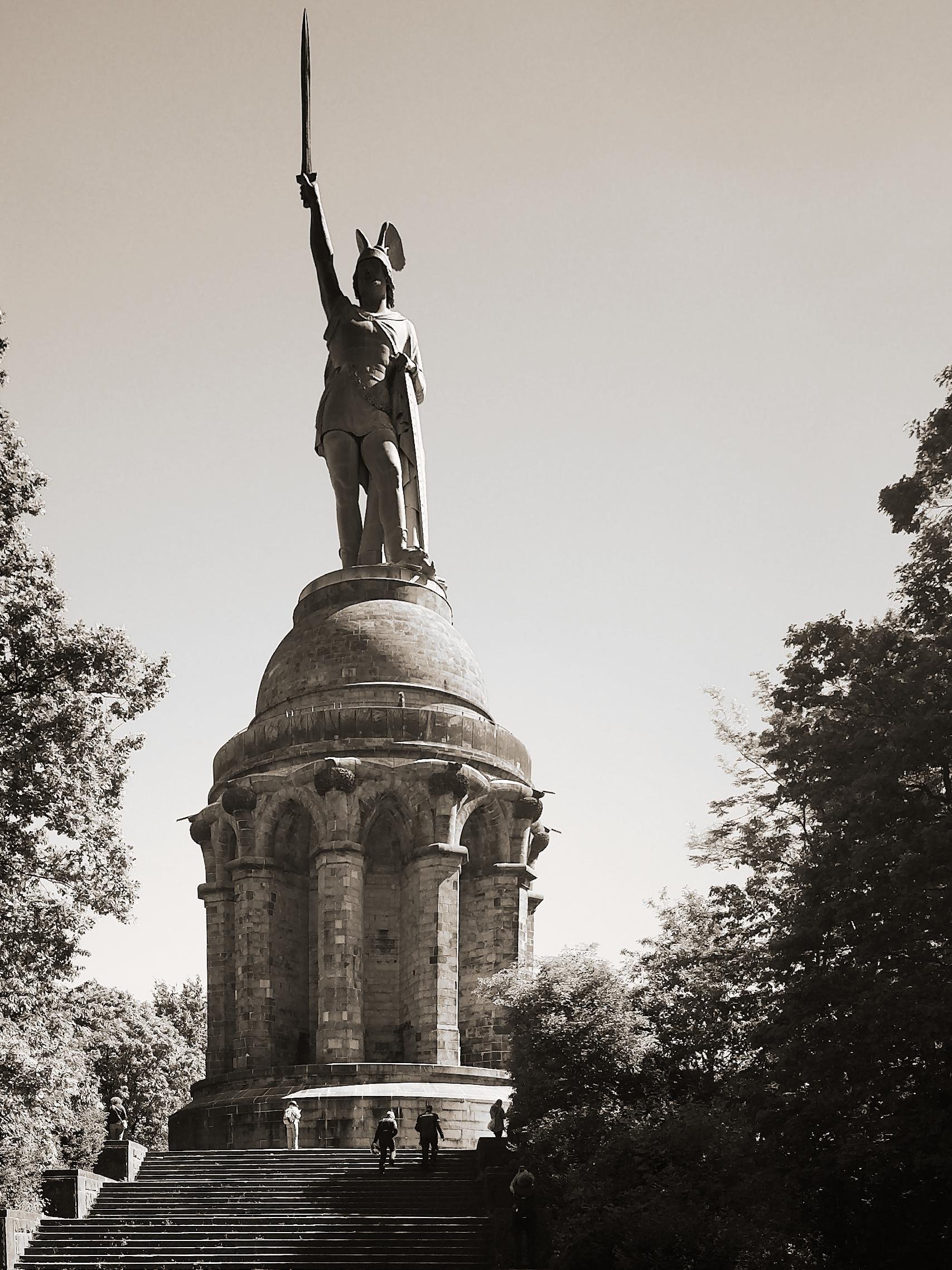 The image shows the Hermannsdenkmal, a monument located in the Teutoburg Forest in Germany. It features a tall statue of Arminius (Hermann), a chieftain of the Cherusci tribe, who is known for defeating three Roman legions in the Battle of the Teutoburg Forest in 9 AD. The statue is mounted on a large, columned pedestal.
