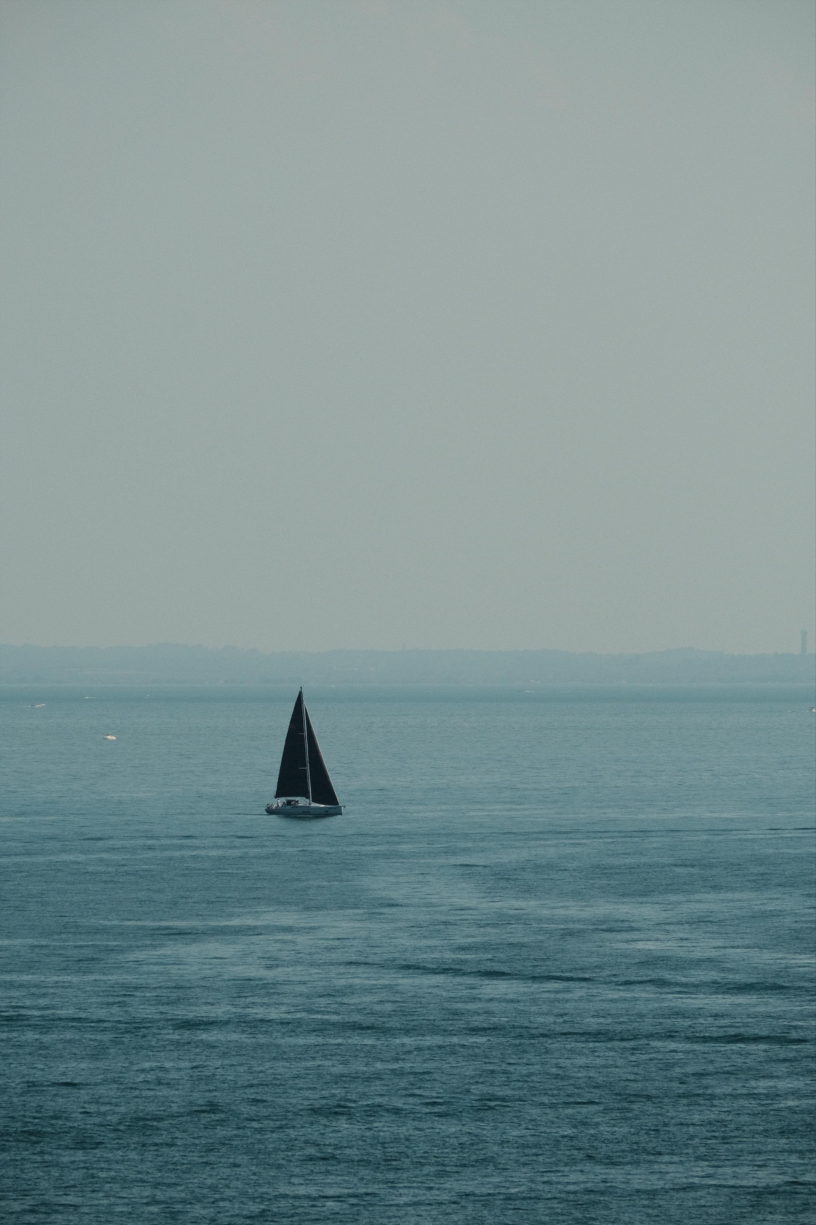 A calm lake with only small ripples by a bit of wind, and a shallow and pale distant coastline in the background under a pale blue sky. A single sailing boat with two black sails across the scene, emphasised by the negative space around it.