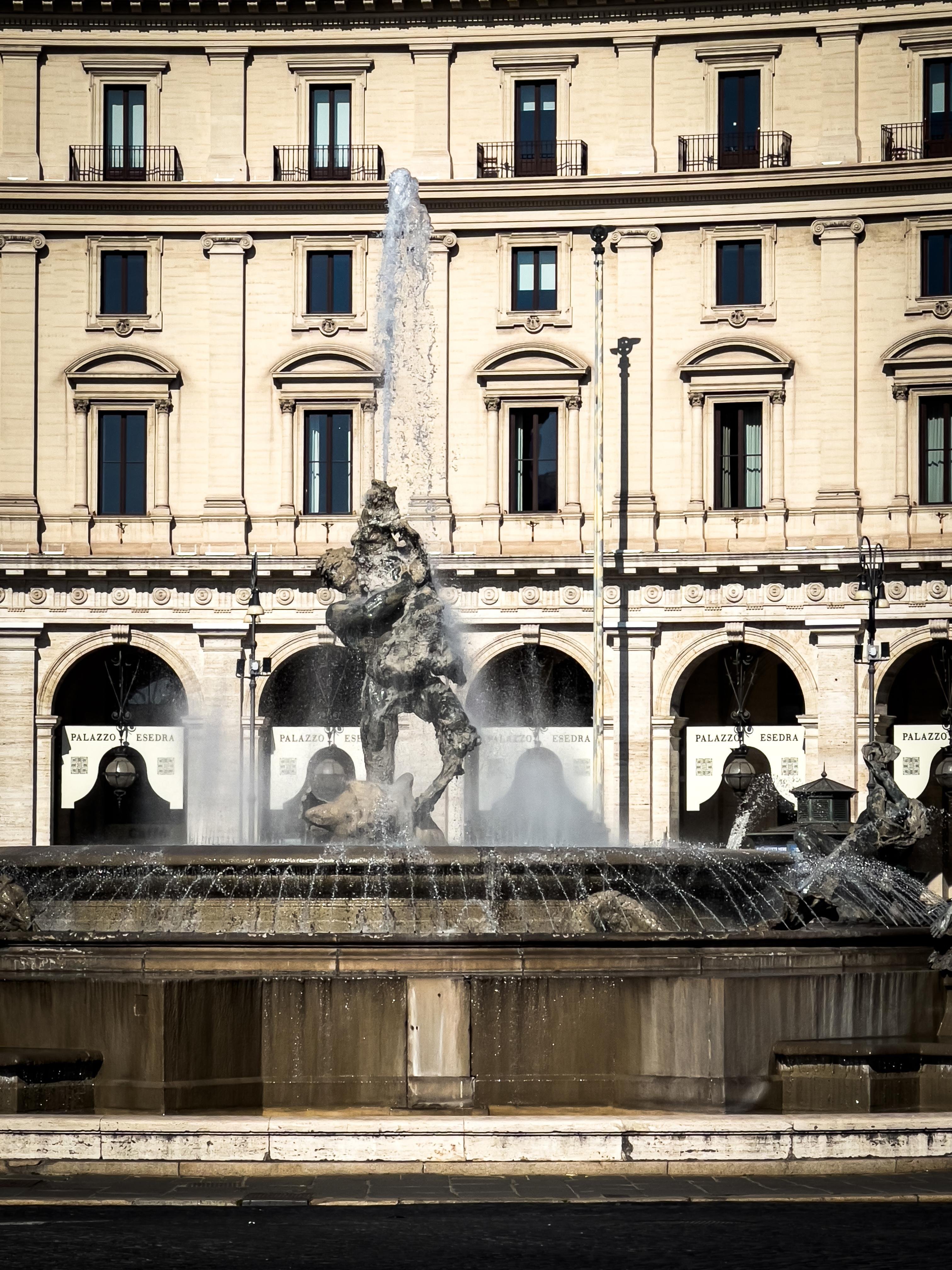 Grande fontana davanti a un edificio storico con più archi e finestre. L'acqua zampilla dal centro della fontana, che è adornata con sculture. L'edificio ha uno stile architettonico classico e sullo sfondo si vedono bandiere con la scritta "PALAZZO".