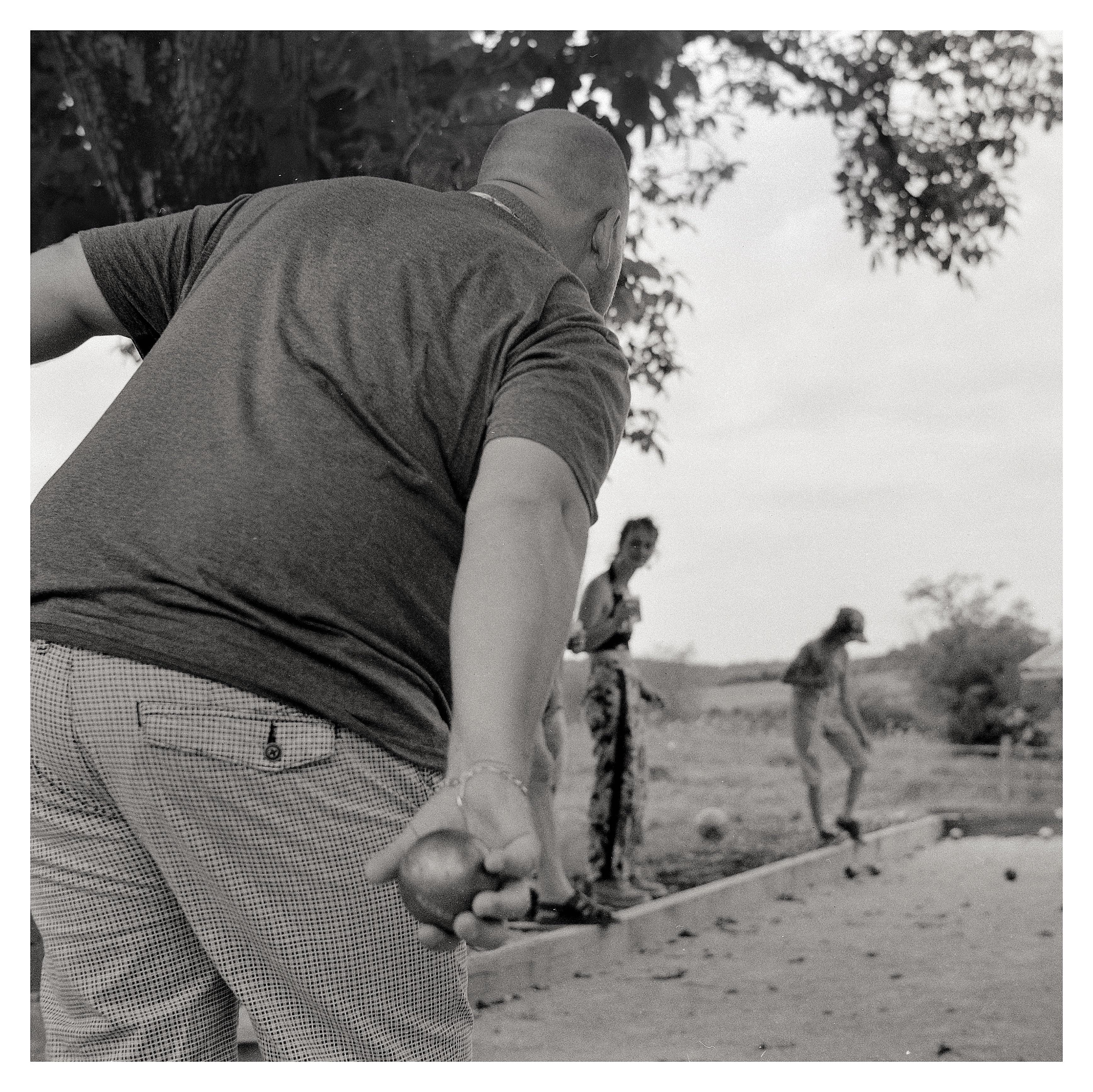 Low-angle black-and-white photo of a man in the foreground holding a metal boule behind his back, preparing to throw on a pétanque court; two other players stand blurred in the background near the far end, with trees and open sky above.