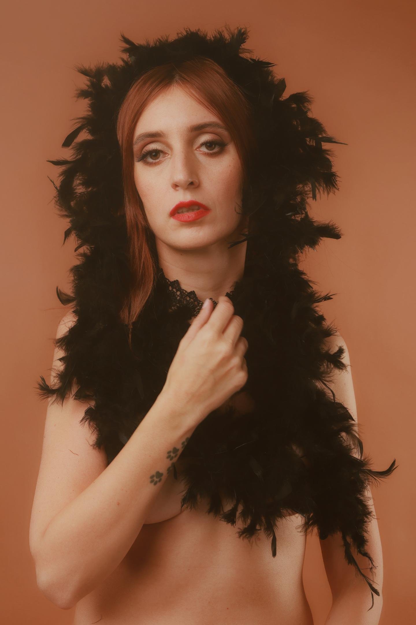 The image shows a person wearing a black feather boa draped around their shoulders and head. They have red lipstick and are posed against a neutral background.