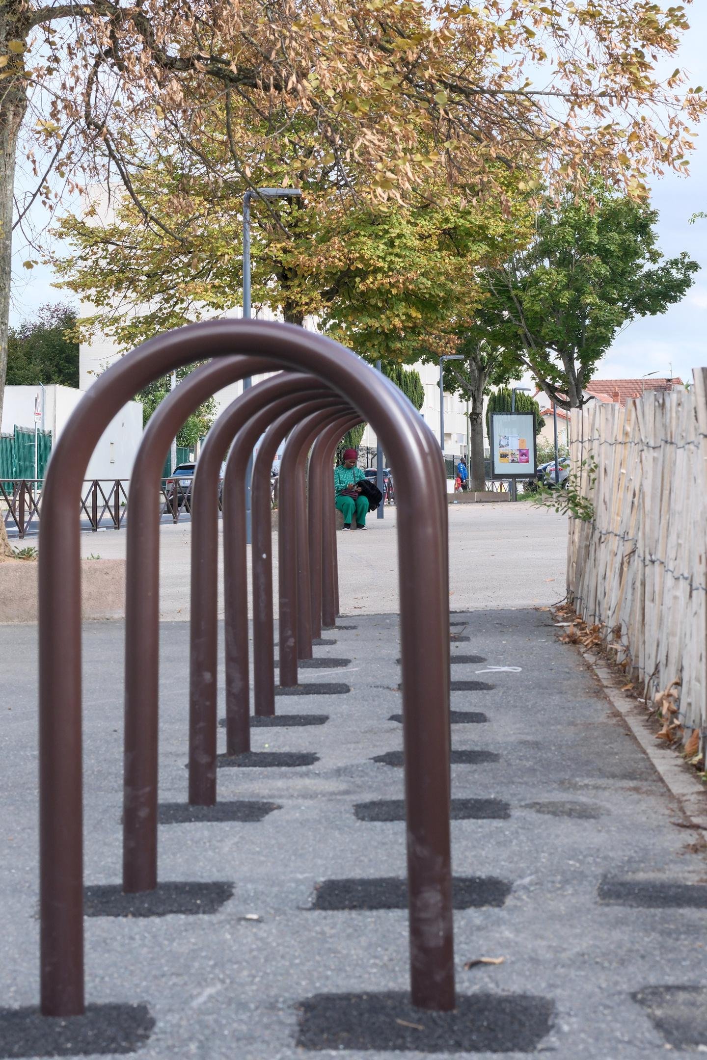 Photo verticale. Prise à hauteur de taille, des arceaux d’attache de vélo les un derrière les autres dirige le regard vers une femme habillée en vert avec un turban pourpre, assise et attendant en regardant son téléphone posé sur sur un sac marron. Une veste noire est posée repliée sur son bras gauche. Un arbre en train de passer aux couleurs d’automne est au dessus d’elle.