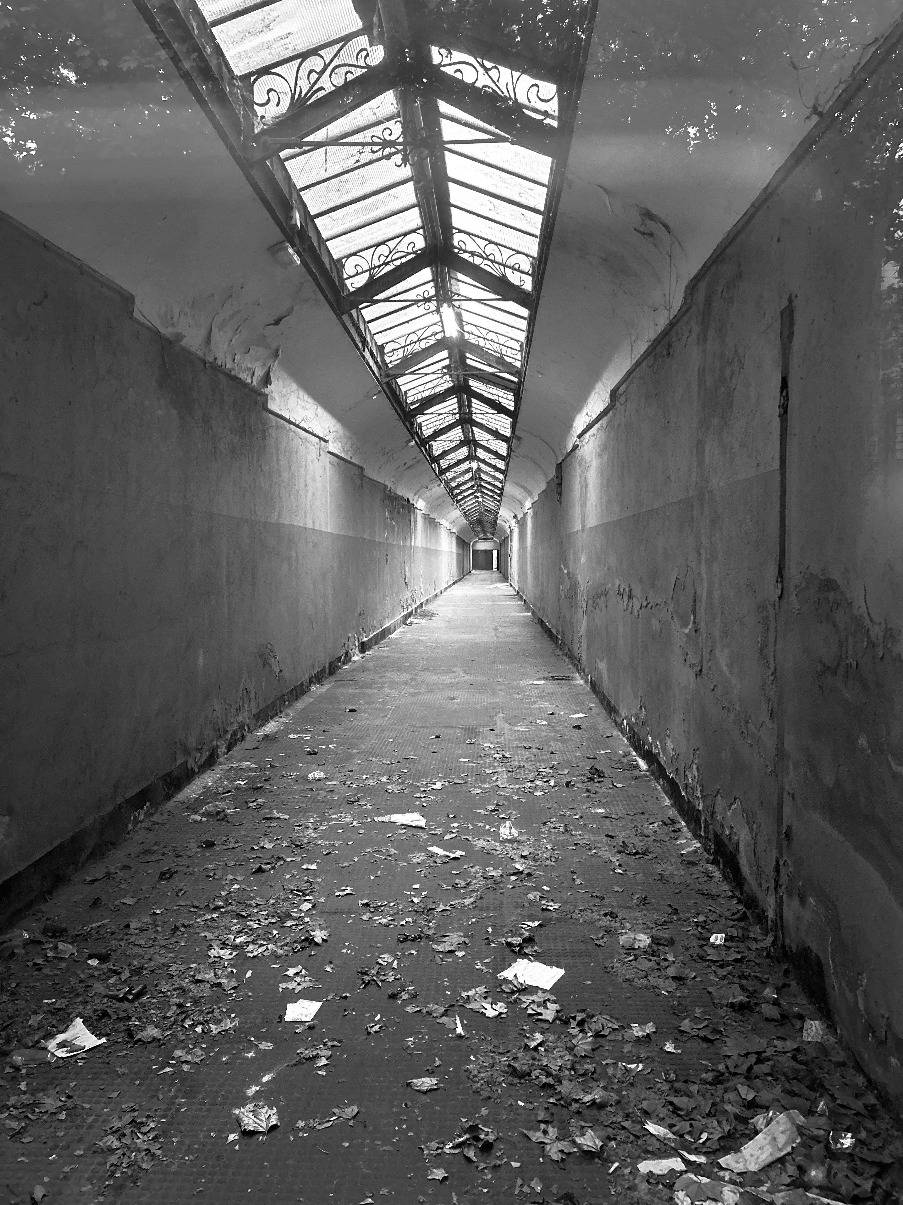 The photo shows a long, empty corridor with a glass ceiling. The floor is covered with leaves and debris, the corridor leads to a doorway in the distance. The image is in black and white.