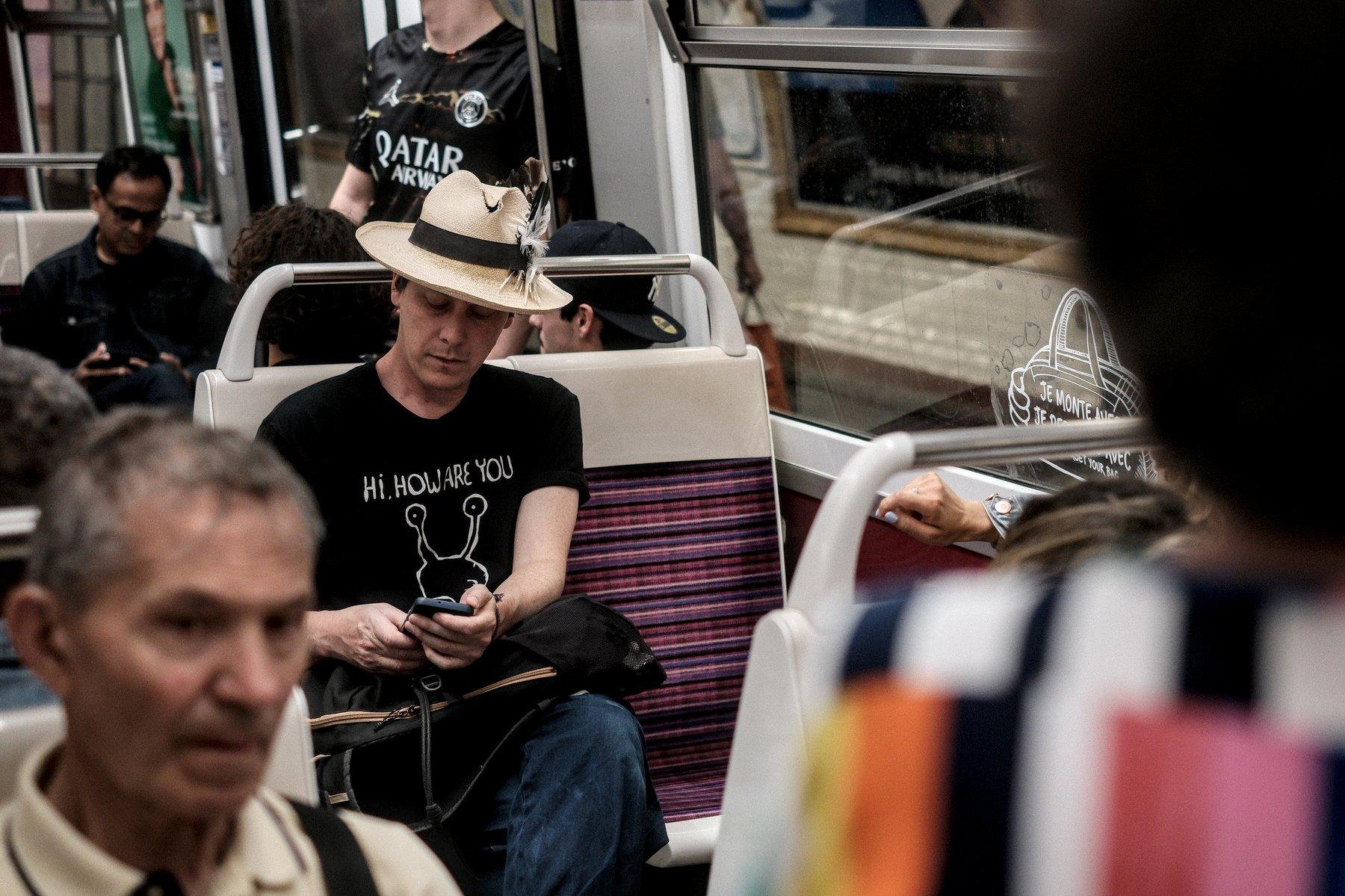 Photo horizontale. Un homme assis dans un carré de 4 dans le métro, en t-shirt noir avec écrit «&nbsp;Hi. How Are You&nbsp;» dessus et une tête d’escargot dessinée. Il porte aussi un jean et un chapeau de paille avec un ruban noir et des plumes accrochées dedans