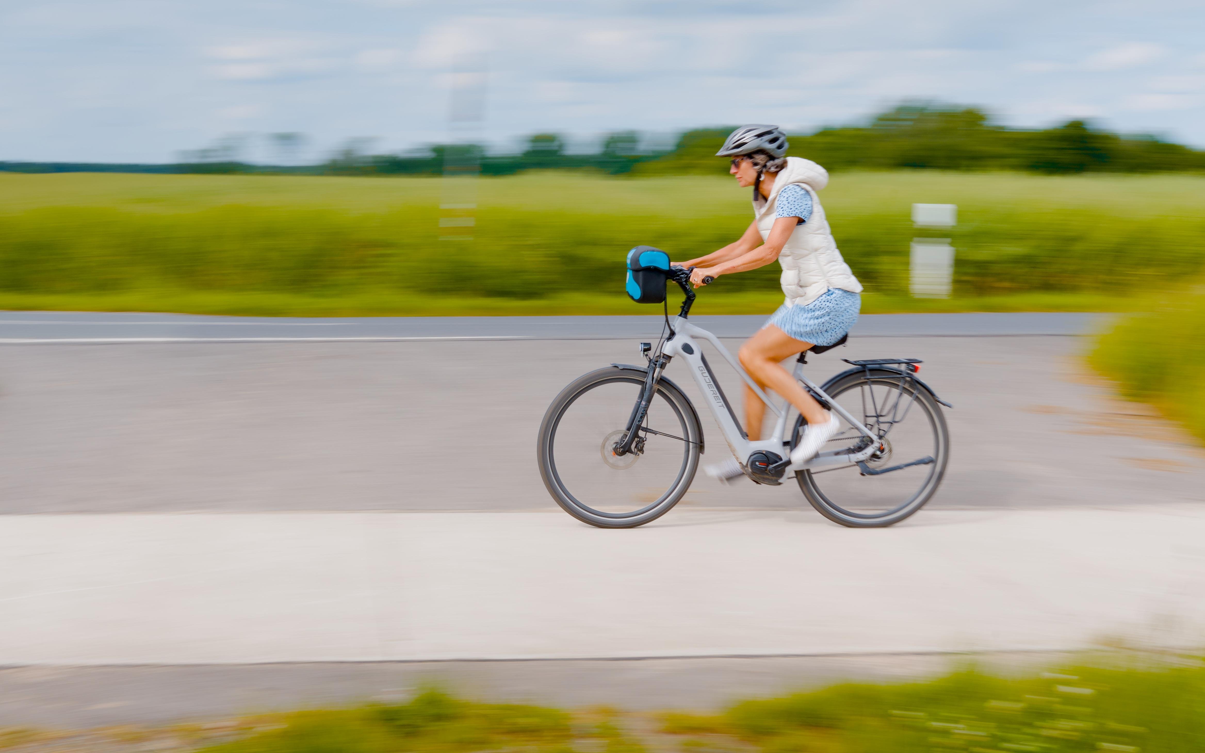 Person wearing a bicycle helmet riding a gray bike with a small blue handlebar bag along a paved road, with motion blur and green fields under a cloudy sky in the background.
###
Eine Person mit Fahrradhelm fährt auf einem grauen Fahrrad mit einer kleinen blauen Lenkertasche eine asphaltierte Straße entlang; im Hintergrund sind – mit Bewegungsunschärfe – grüne Felder unter einem bewölkten Himmel zu sehen.