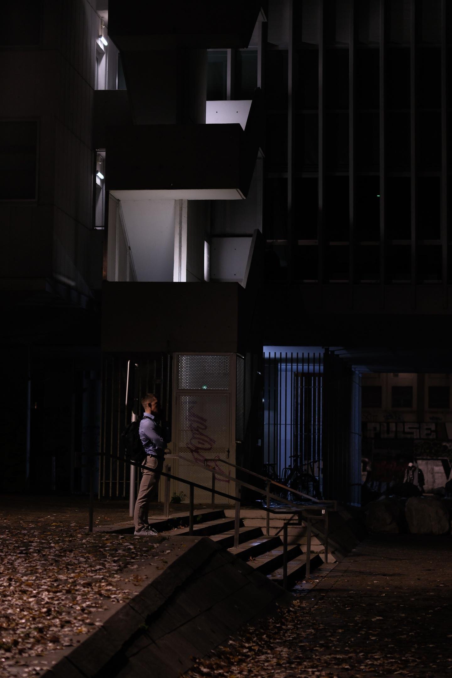 Photo de nuit au format portrait d’un homme de profil, en chemise en haut d’une petite volée de marches, devant un immeuble dont on voir la cage d’escalier éclairée de lumières blafardes. Une lumière plus chaude arrive depuis le coin inférieur gauche de la photo, le reste est plongé dans le noir