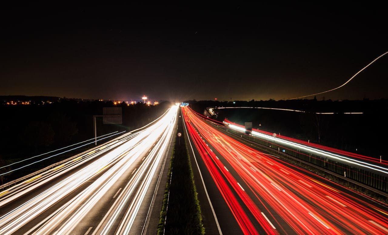 This image shows a long-exposure photograph of a highway at night. The white and red light trails represent the headlights and taillights of vehicles moving along the road. The bright trails create a dynamic effect against the dark background, capturing the motion and speed of the traffic.