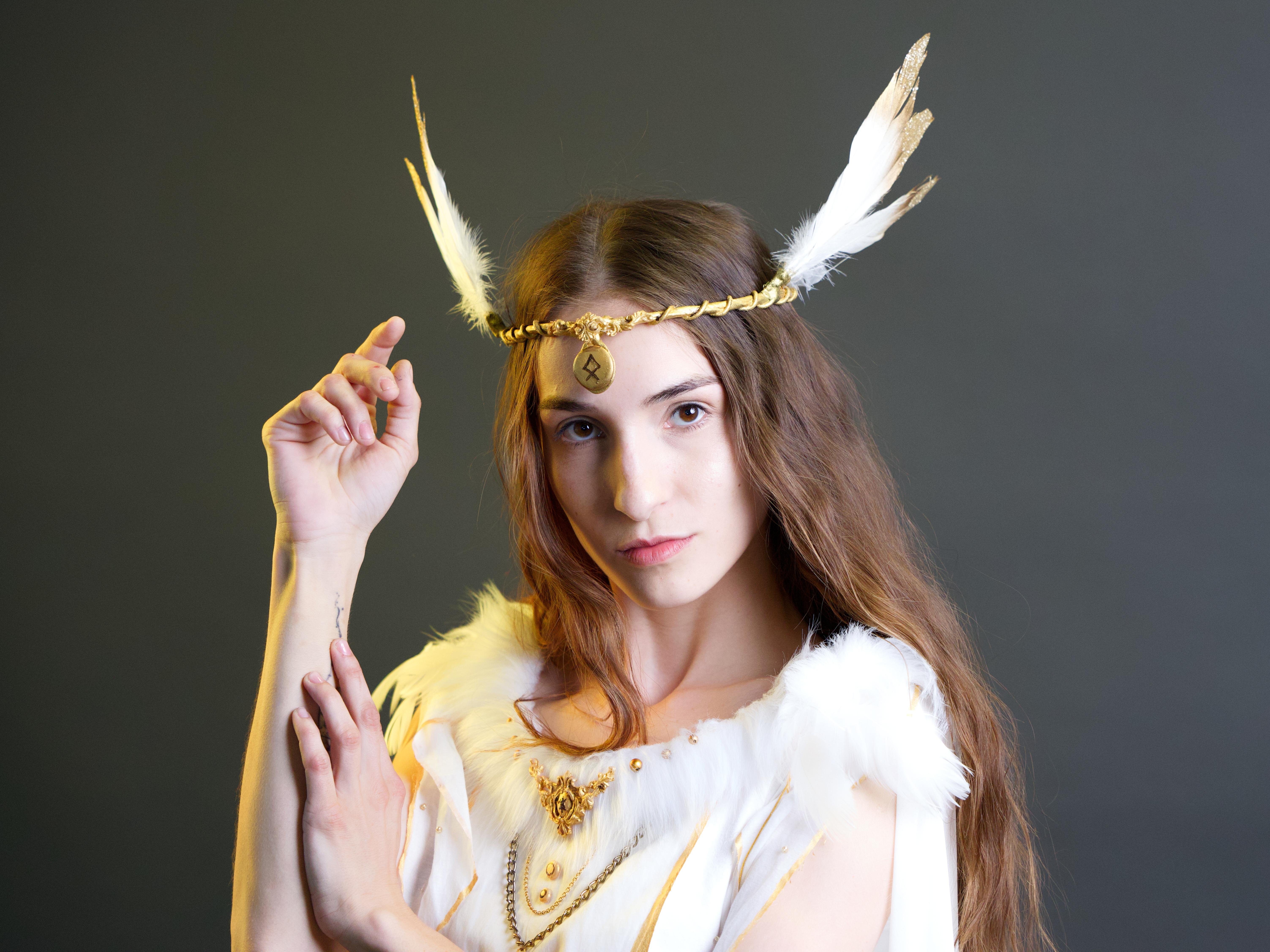 The image shows a person dressed in a costume that appears to be inspired by historical or fantasy themes. They are wearing a headband with feathers and a garment adorned with feathers and decorative elements. The pose and styling suggest a portrayal of a mythical or legendary character.