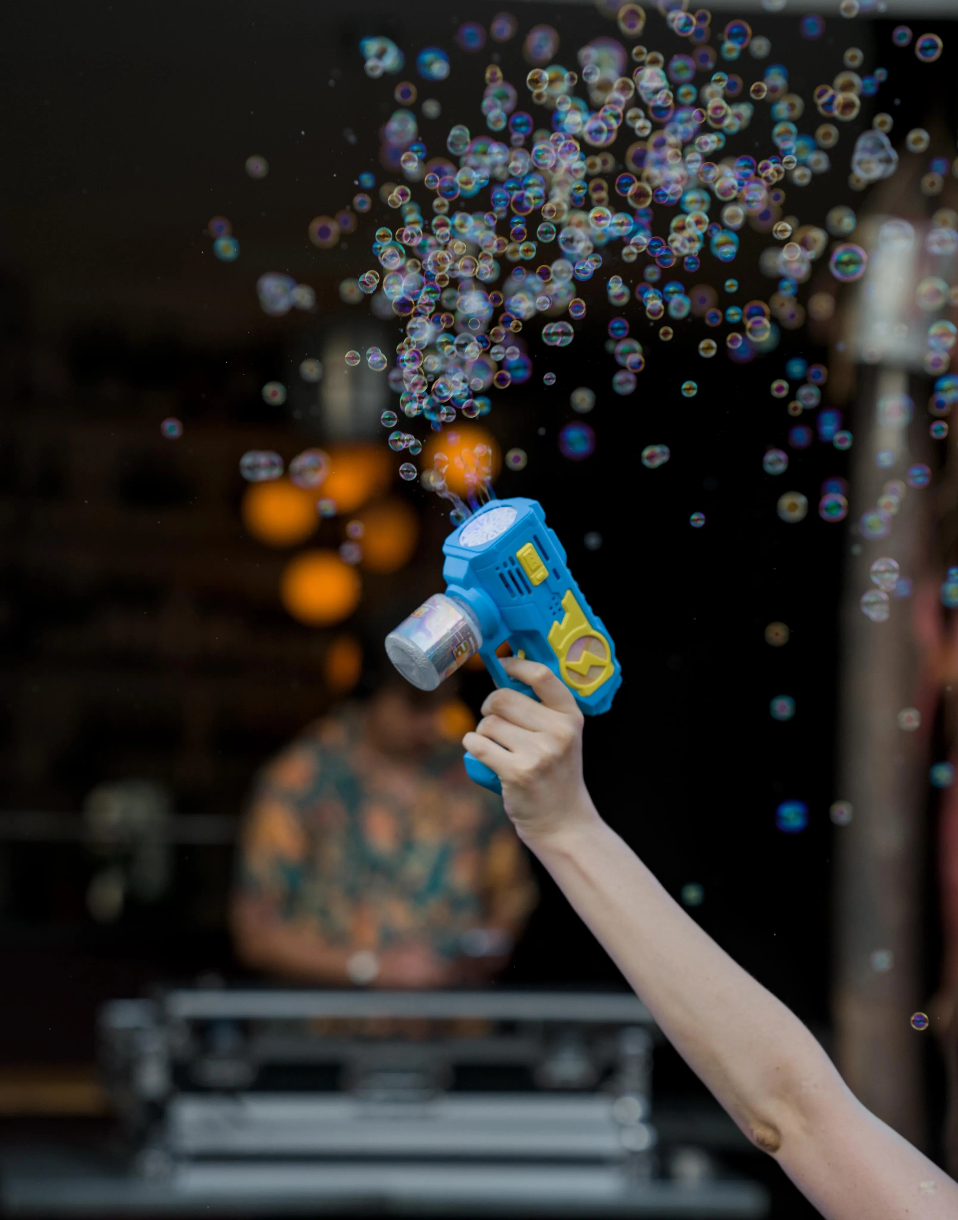 Photograph of a bubble gun, held by the extended arm of an unseen person. The bubble gun has a blocky shape and is painted bright blue and yellow. Many bright, small soap bubbles are floating in the air above and around the gun. The gun is also producing new bubbles. In the background out-of-focus blur, a DJ with a green and white shirt is mixing music.