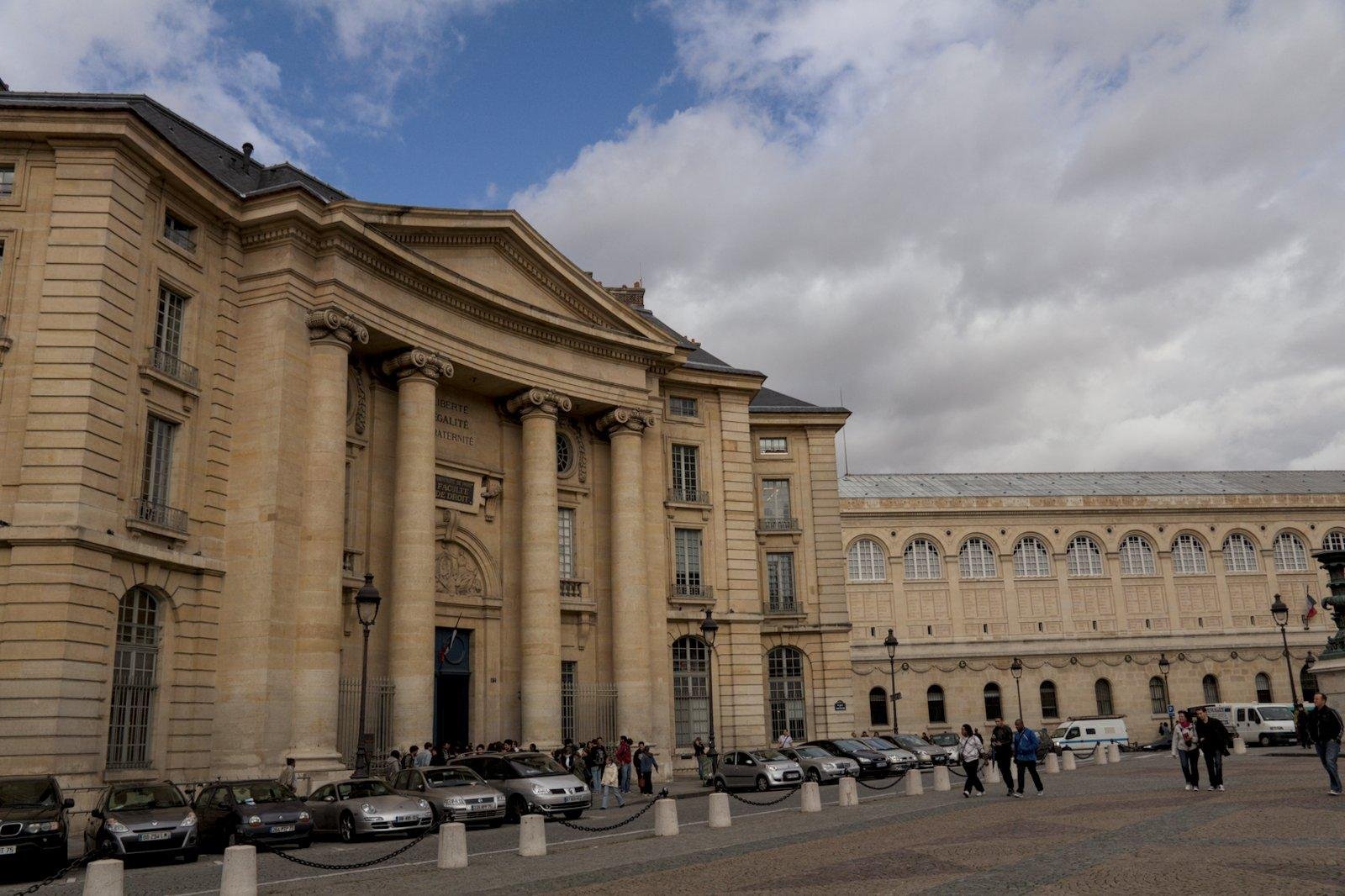 Photo format paysage montrant un bâtiment avec une façade rappelant l’architecture grecque, avec des colonnes et le fronton. Des voitures sont garées devant. Il s’agit de la faculté de droit de Paris