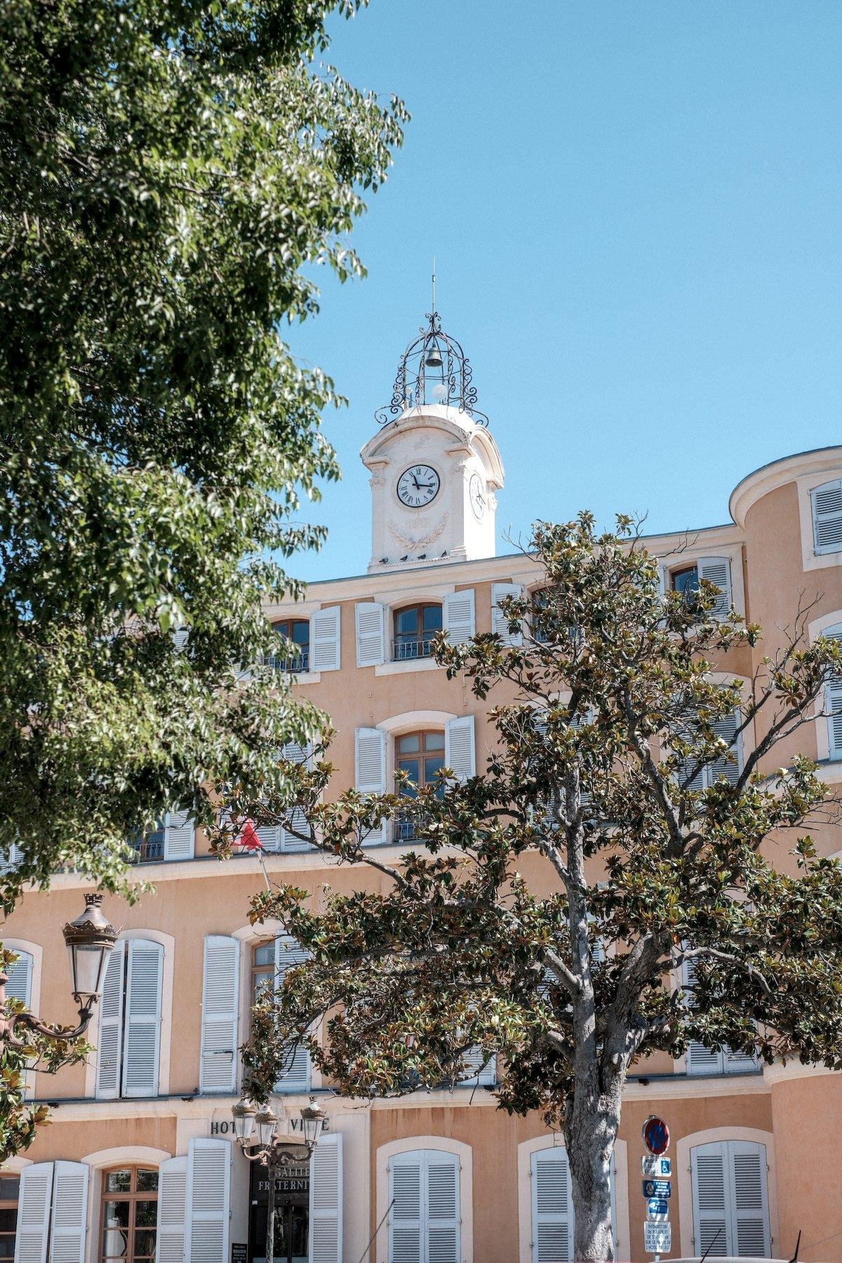 Photo verticale. Derrière deux arbres, un bâtiment ocre avec des fenêtres aux volets blancs, surmonté d’une petite tour avec une horloge et dessus, une cloche dans une armature en fer forgé, le tout sous un ciel bleu