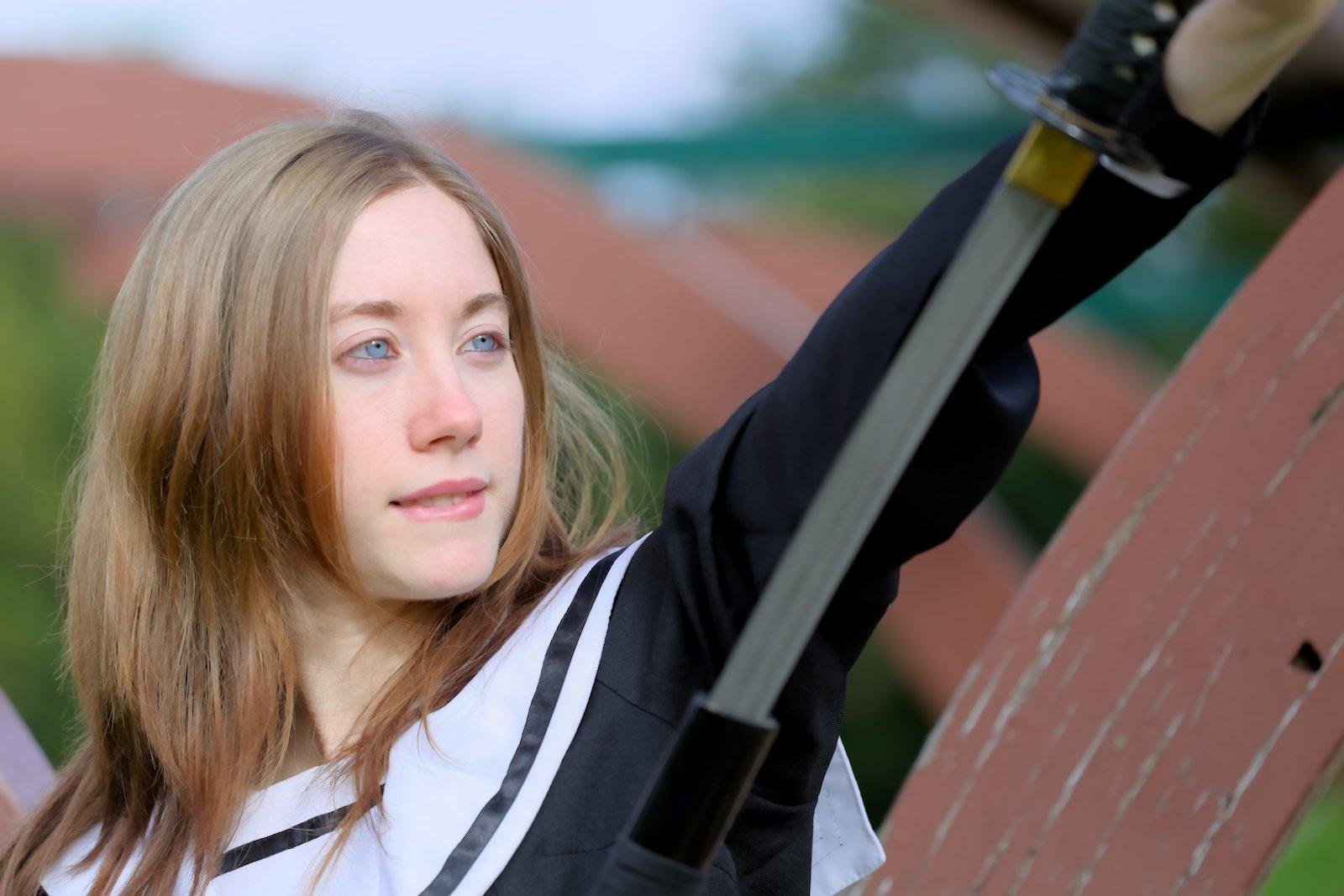 Photo format paysage d’Une jeune femme aux cheveux roux, long aux épaule, qui dégaine un katana, en le regardant avec avidité