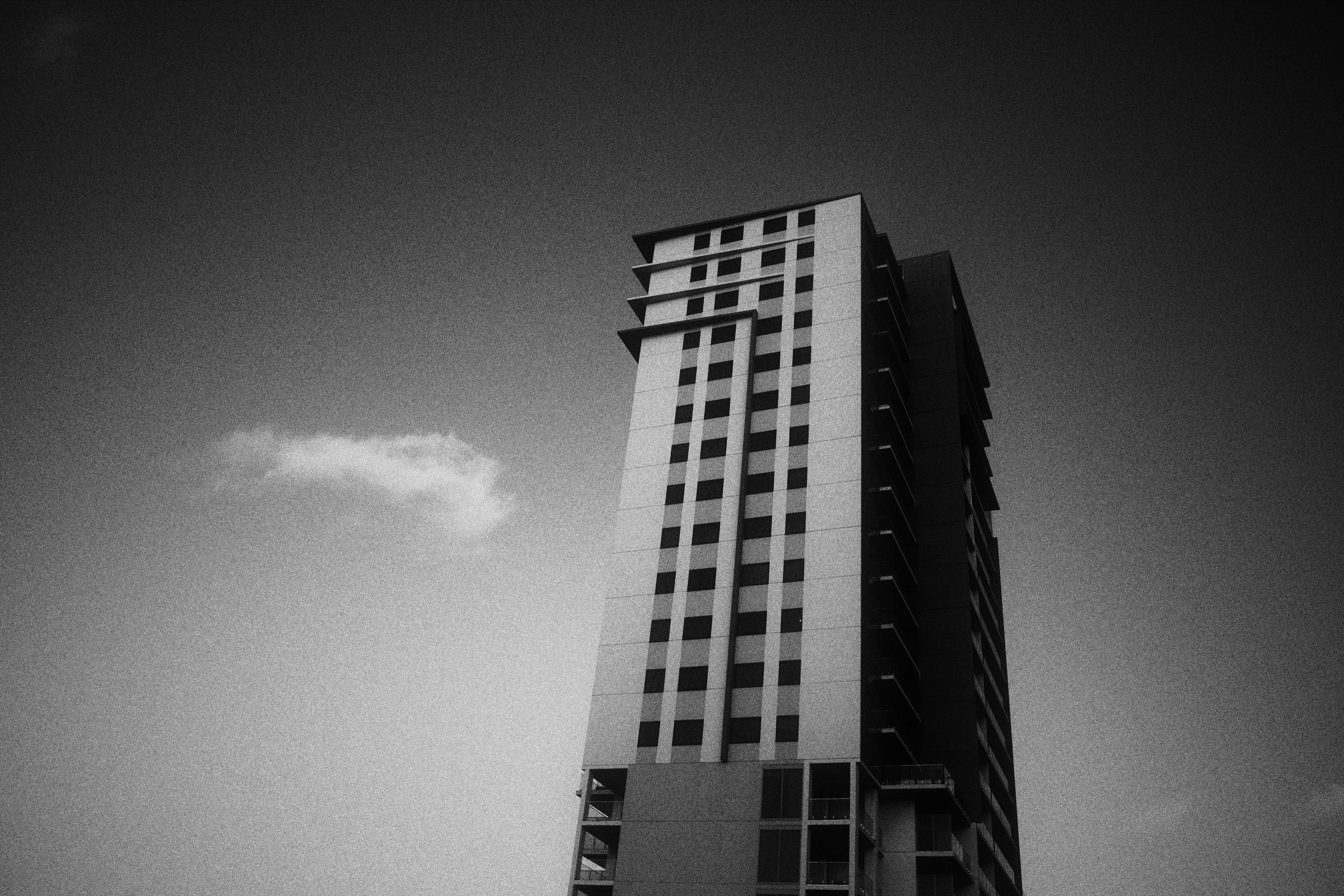 A tall, rectangular building with multiple floors and windows under a dark sky with a cloud on the left. Other sparser clouds are visible in the frame but don’t stand out as much.