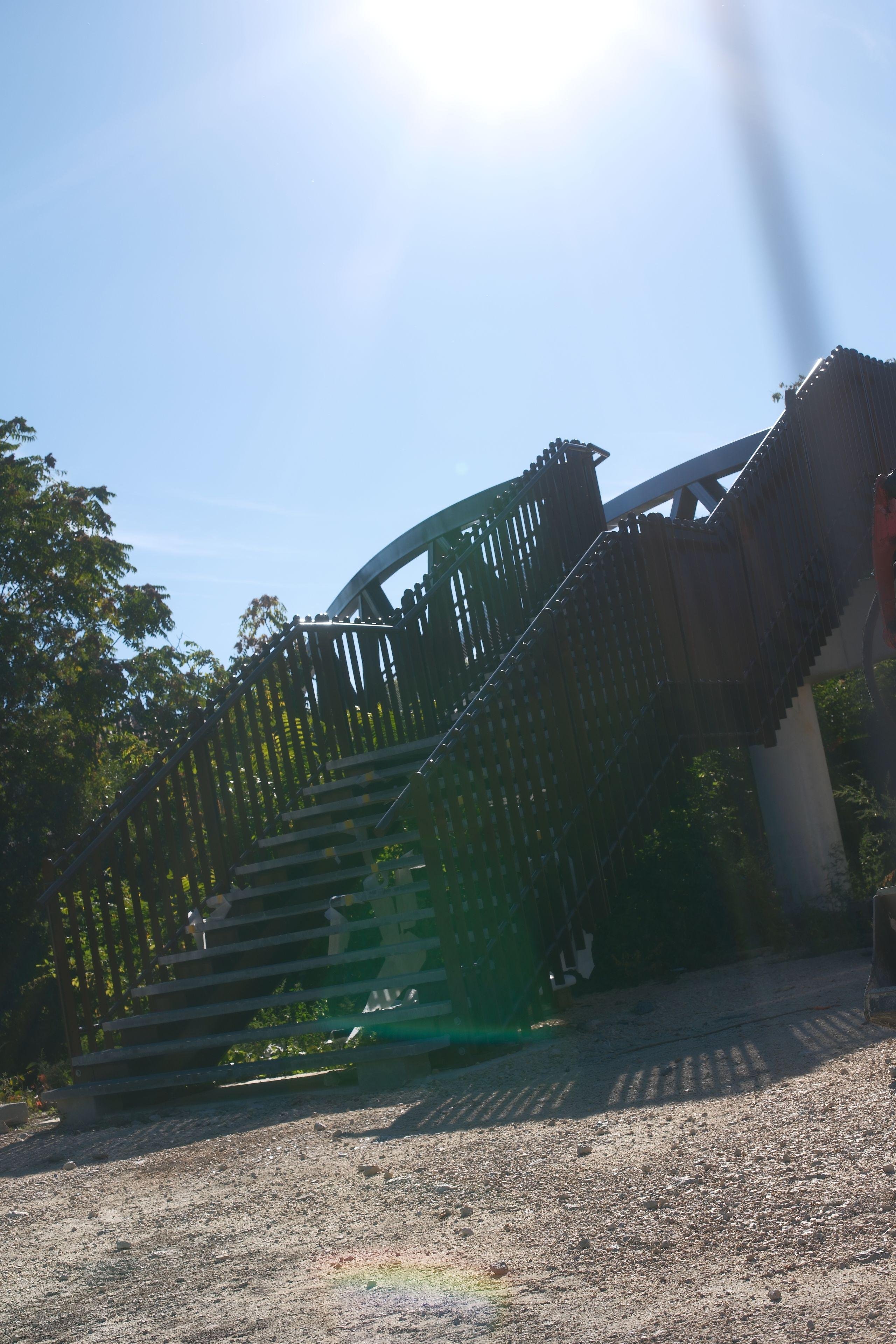 Photo format portrait, montrant un escalier extérieur de biais, avec des barrières de chaque côté, menant à un pont pour piéton dont on ne voit que 2 bouts de structure, éclairé par le soleil en haut de l’image, formant un flare vert sur le bas de l’image. Le sol est de la terre battue et des cailloux