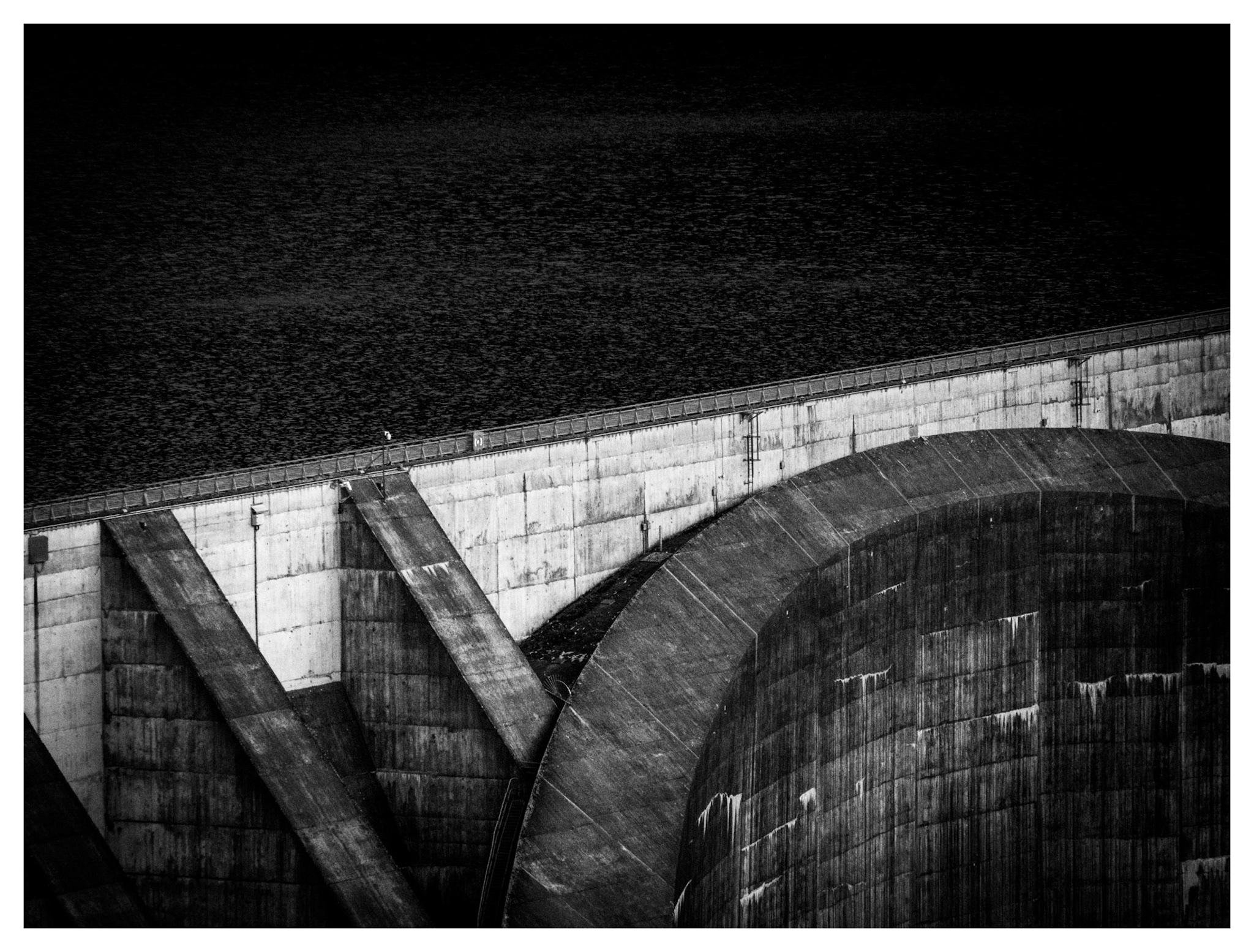 Black-and-white photo of a concrete dam viewed at an angle, with dark water behind it, a curved spillway wall in the foreground, and diagonal support buttresses along the dam face.