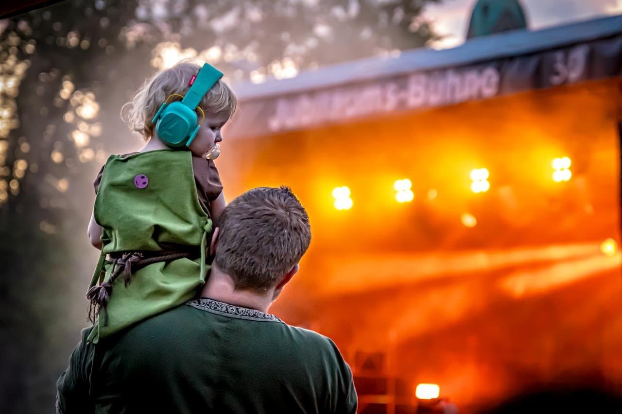A child wearing large teal earmuffs sits on an adult's shoulders facing a stage. The child is dressed in a green outfit with a small purple patch on the back. The stage is illuminated with bright yellow-orange lights, creating a warm glow and silhouetting the child and adult. Trees and a partly visible sign are in the background.