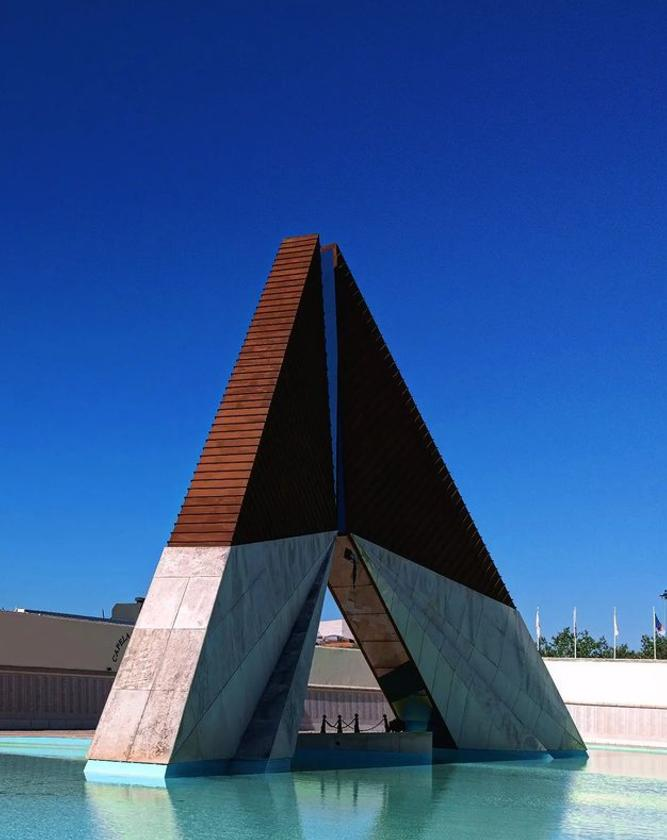 The image shows the Monumento aos Combatentes do Ultramar, a war memorial in Lisbon, Portugal. It features two large triangular structures meeting at the top, situated in a pool of water. The background is a clear blue sky.