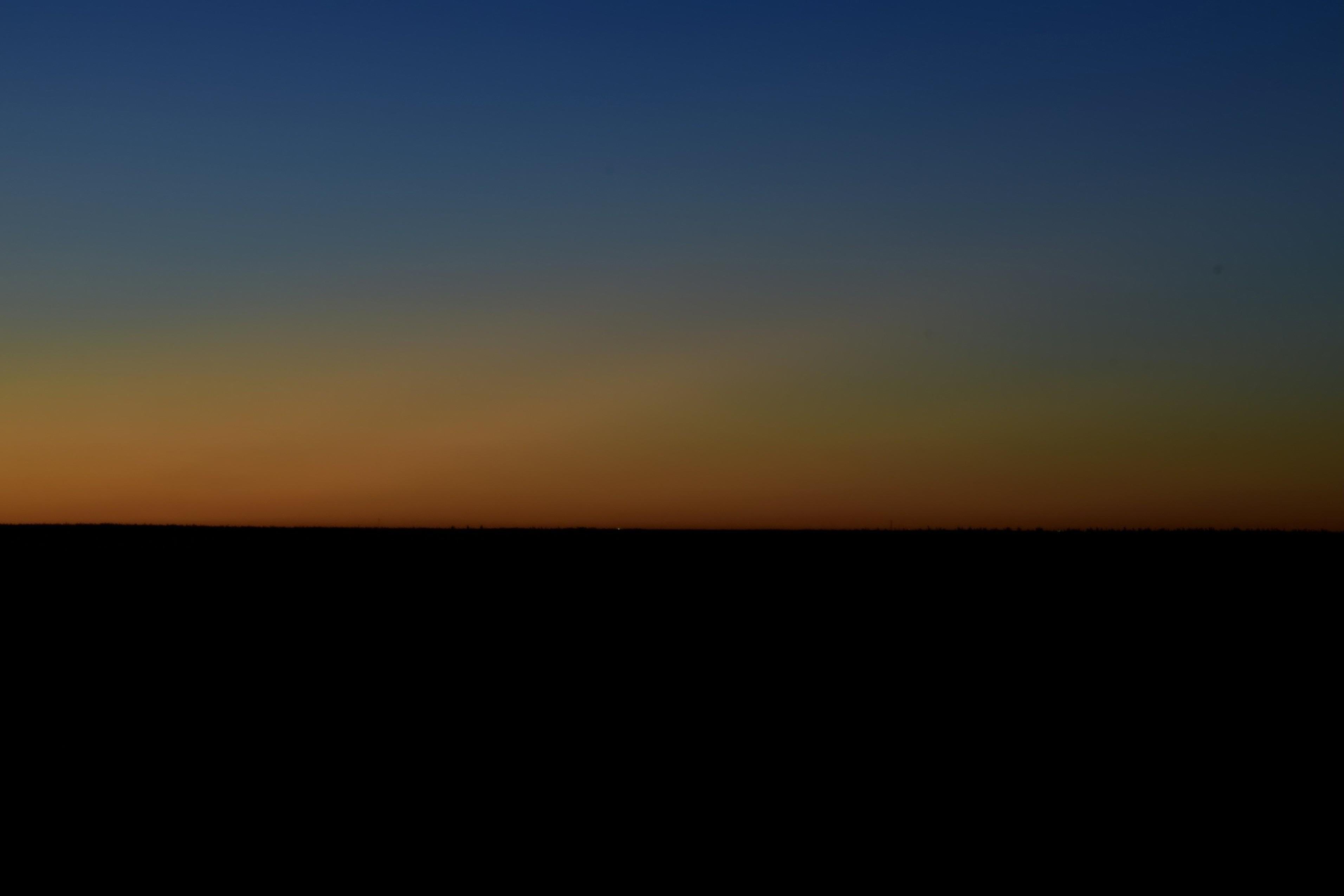 A colorful gradient in the sky, deepening blue up high fading into reds and oranages below. Shades of green and yellow show on the right, further away from the set sun below deeper hues of blue. A dark featureless horizon stretches out underneath