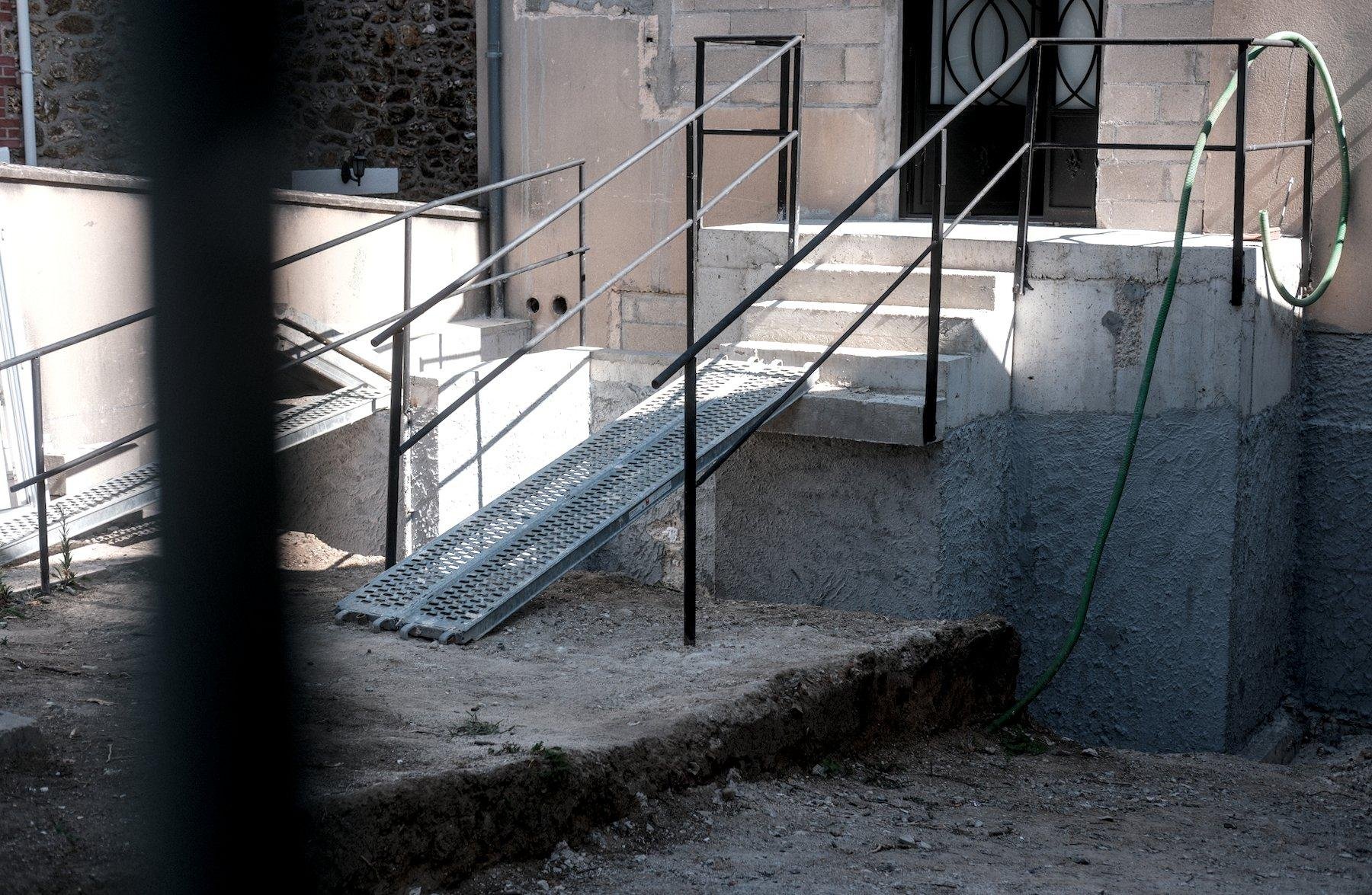 Photo horizontale. À travers une grille, un chantier en cours avec un escalier qui s’arrête à mi hauteur, complété par une double rampe en acier
