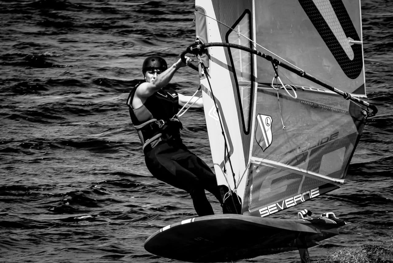 Black-and-white close-up of a windsurfer wearing a helmet and life vest, leaning back while holding the boom on a sailboard cutting across choppy water; the sail fills the right side of the frame.