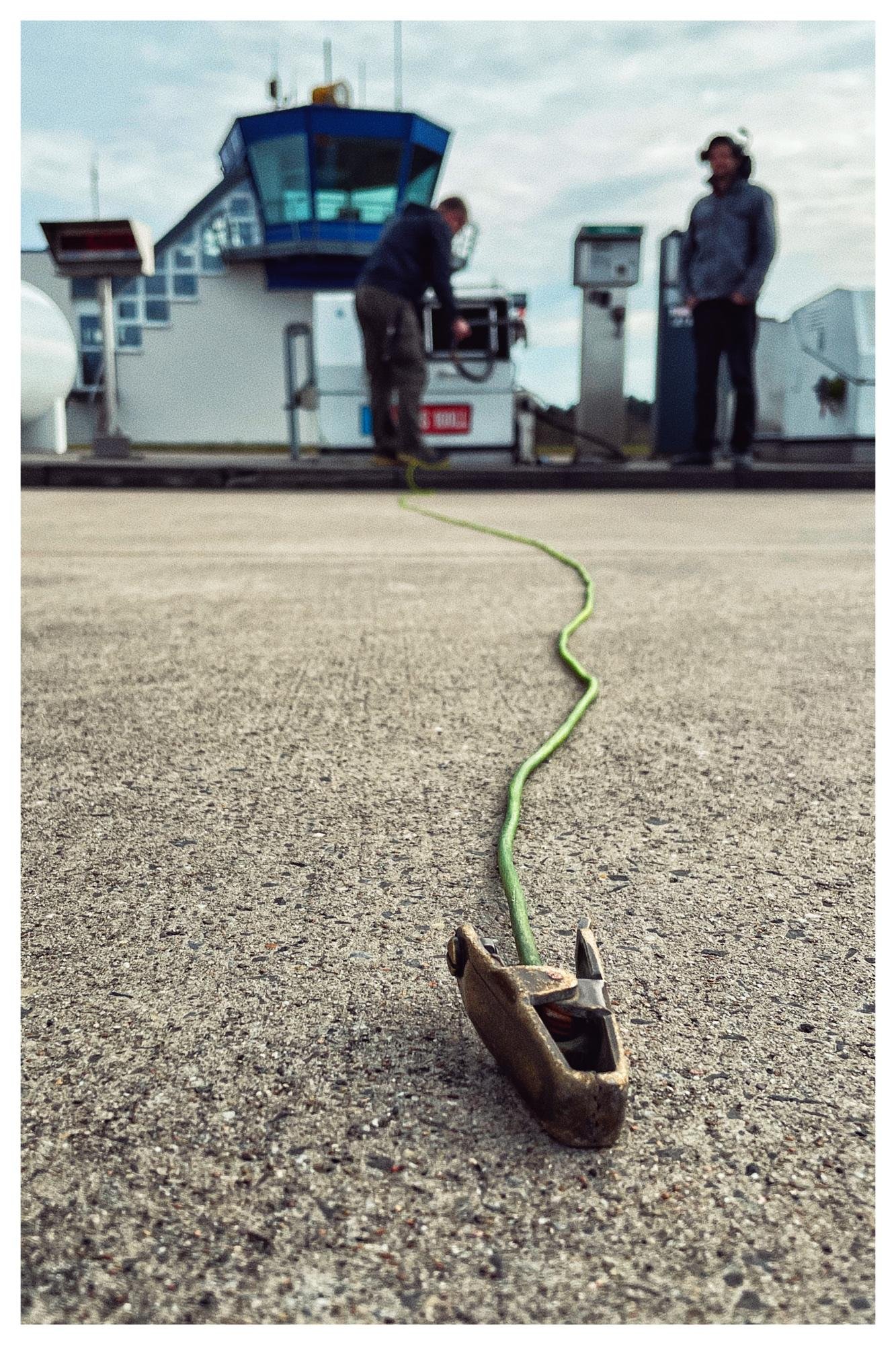 Metal clamp attached to a green cable on an airport tarmac in the foreground, with two ground crew near equipment and an air traffic control tower blurred in the background.