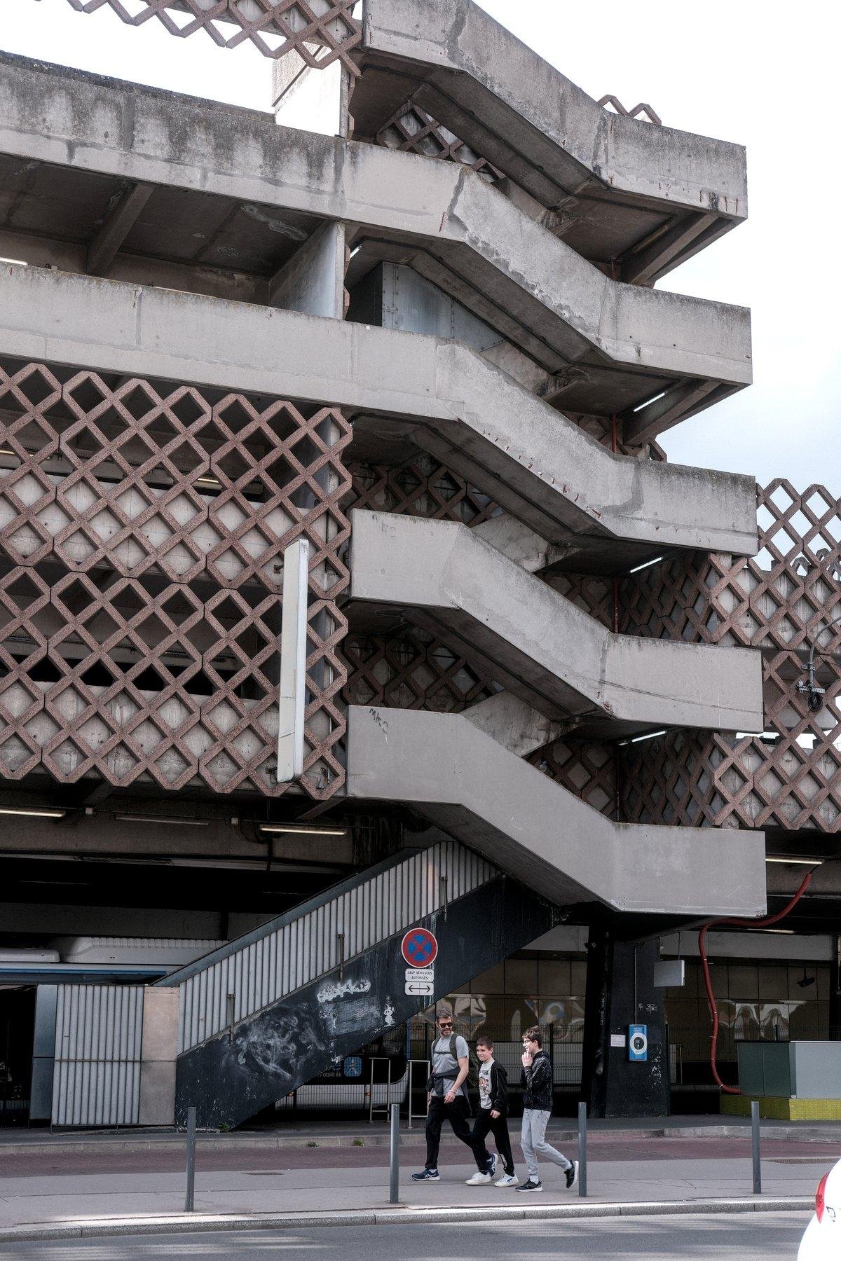 Photo verticale. Des escaliers extérieur en béton gris desservant un bâtiment de 5 étages. 3 personnes marchent sur le trottoir vers la gauche