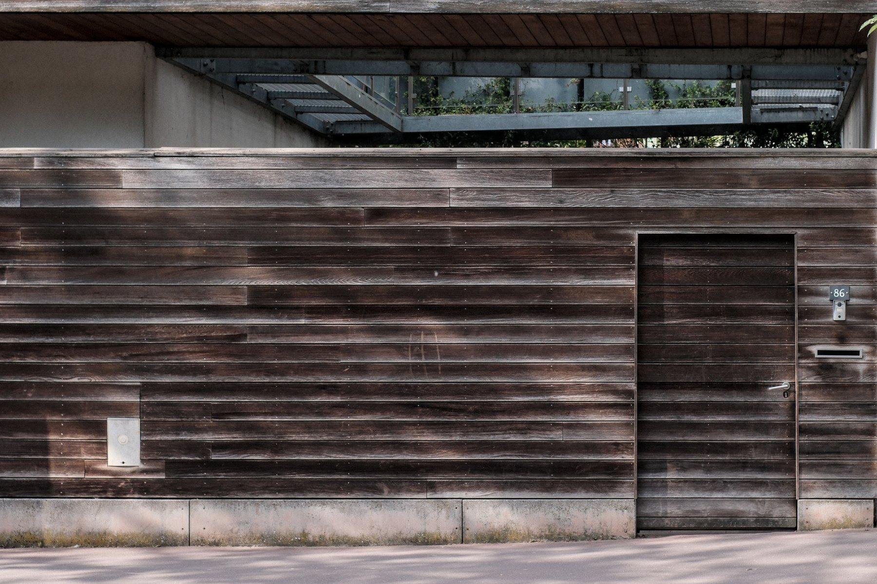 Photo horizontale. Façade de bâtiment couverte de lambris marron sombre et usé par les éléments. Une porte avec le même lambris est visible et semble tenter de se fondre dans le décor