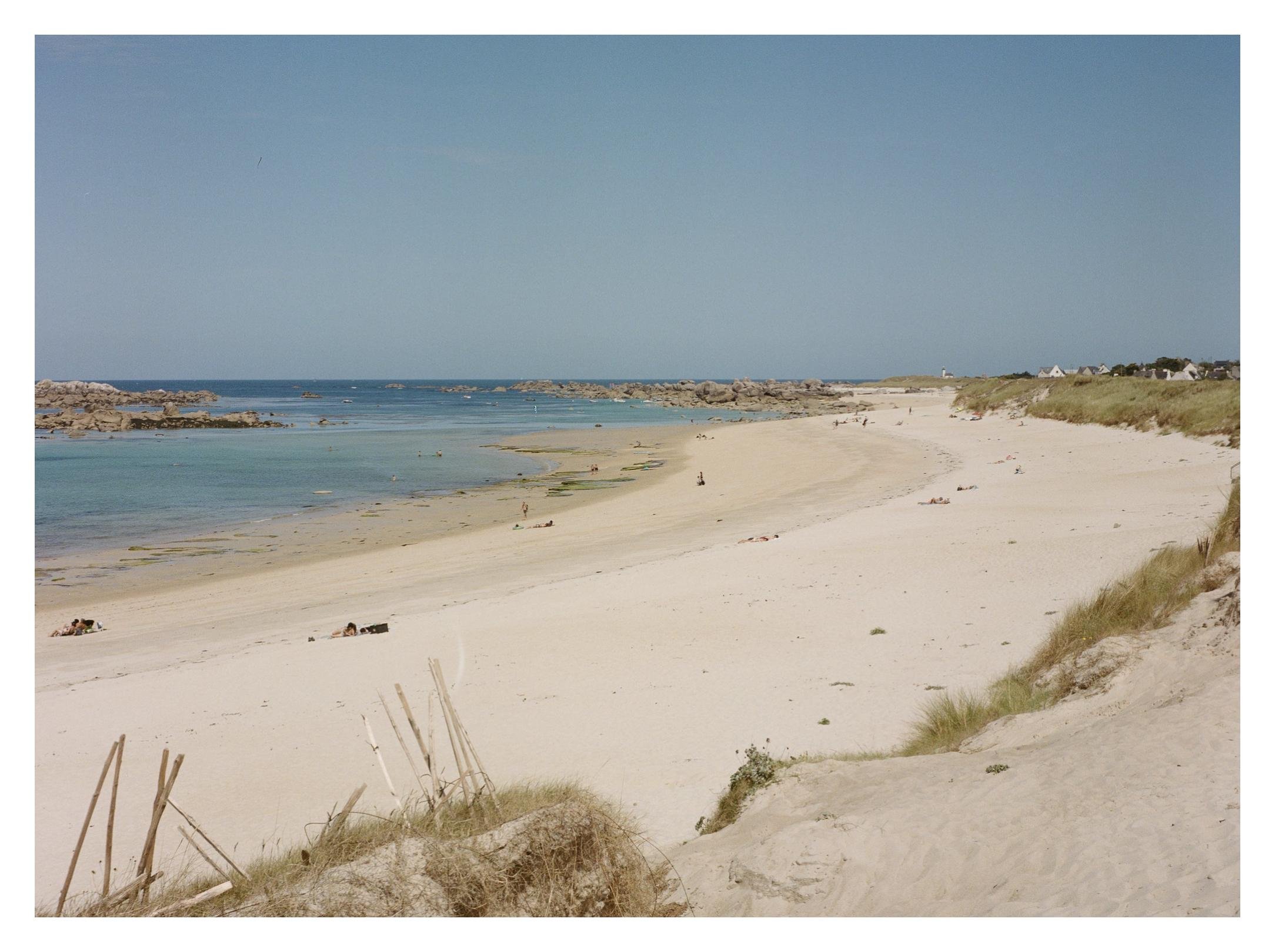 Wide sandy beach curving along a calm blue shoreline, with low dunes and beach grass in the foreground, rocky outcrops in the water, a few small figures scattered on the sand, and distant houses under a clear sky.