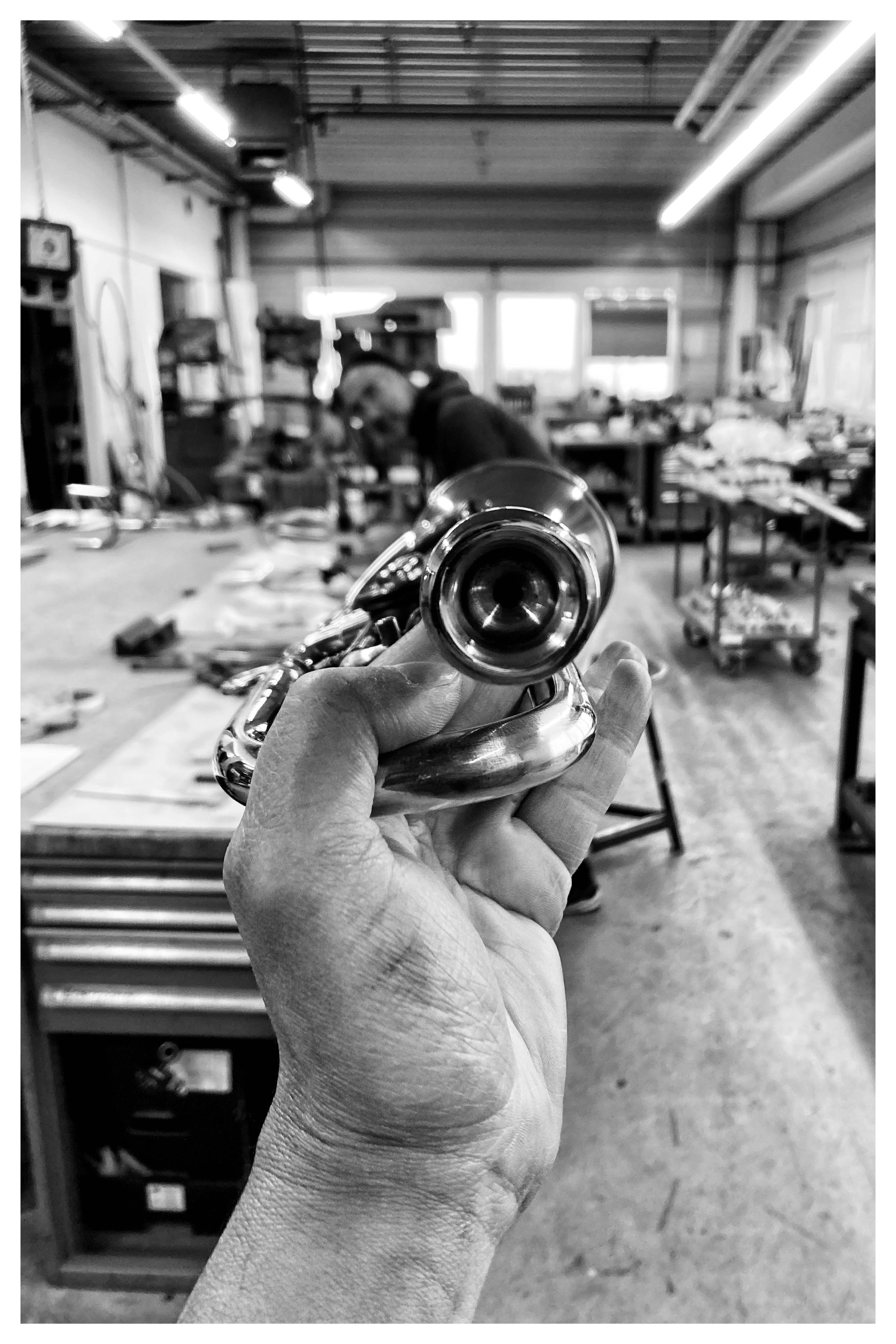 Hand holding a French horn mouthpiece in the foreground. A workshop with various tools and equipment fills the background. Black and white image with a person working at a bench, blurred due to shallow depth of field. Bright overhead lights illuminate the space.