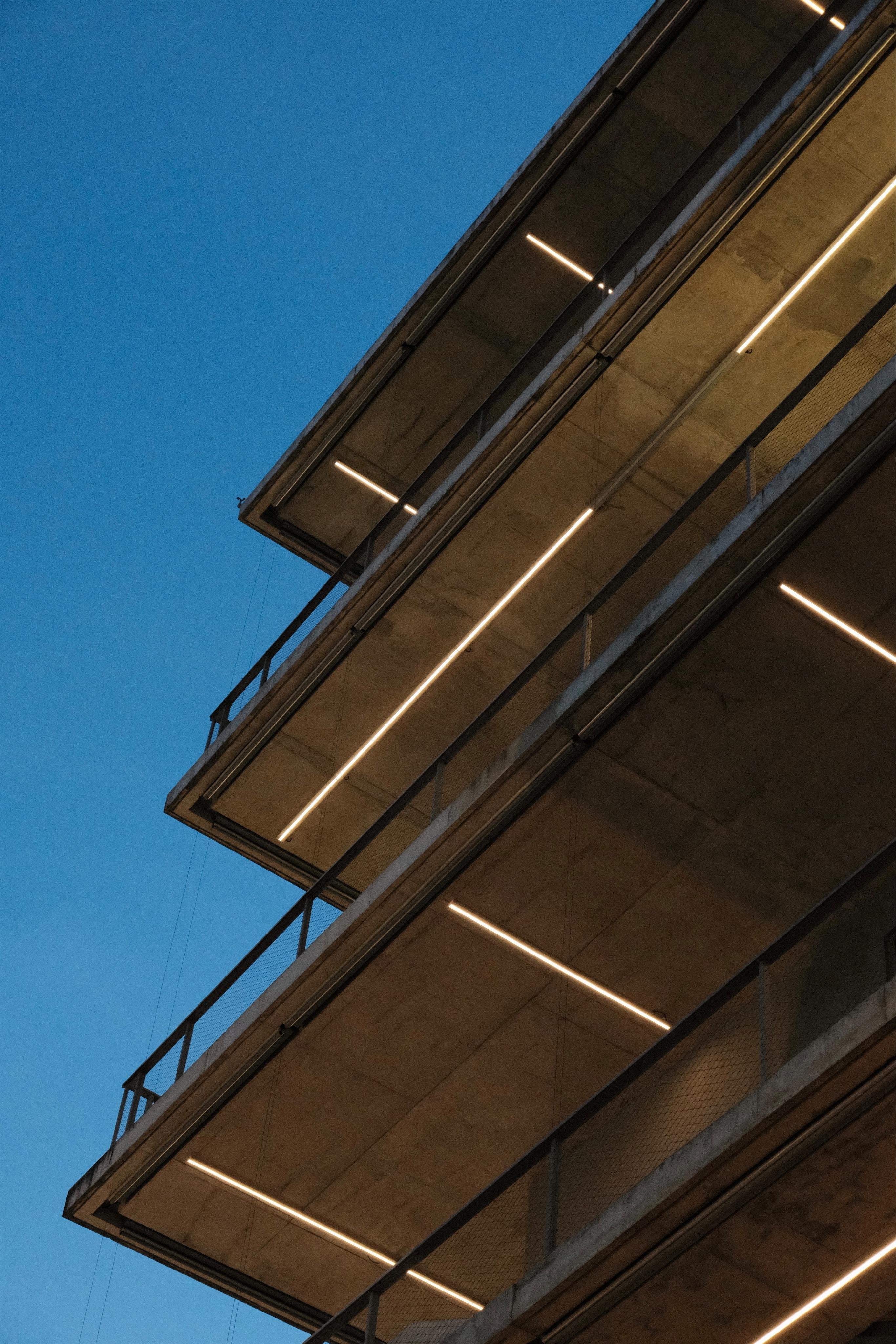 Low angle view of a concrete building with balconies running all along the building. What you can see is the undersides of these balconies of three stories „stacked“ behind each other. The facade is not visible due to the steep angle. On the visible surfaces, alternating patterns of white neon light tubes are attached to illuminate the walkways on the balconies.
In the background a deep blue sky short after sunset.