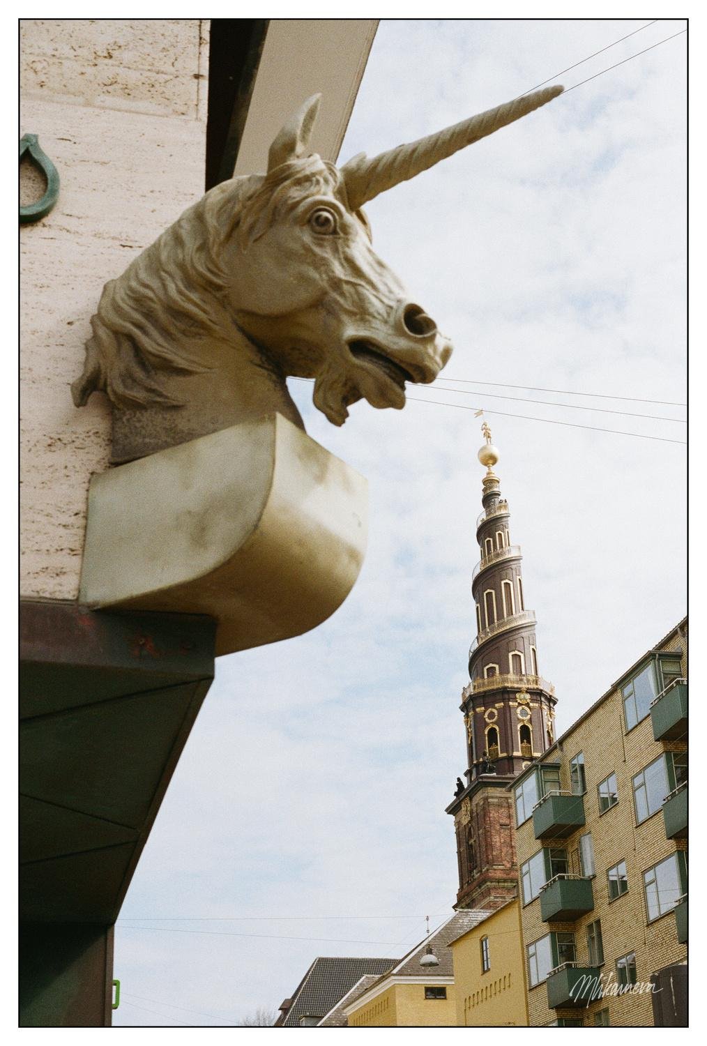 Stone unicorn bust mounted on a building corner with its head facing right. In the background, a tall, ornate tower with gold accents and a spiral staircase is visible against a partly cloudy sky. Below the tower, there are yellow and brick buildings with multiple windows and balconies. Thin wires are strung across the sky.