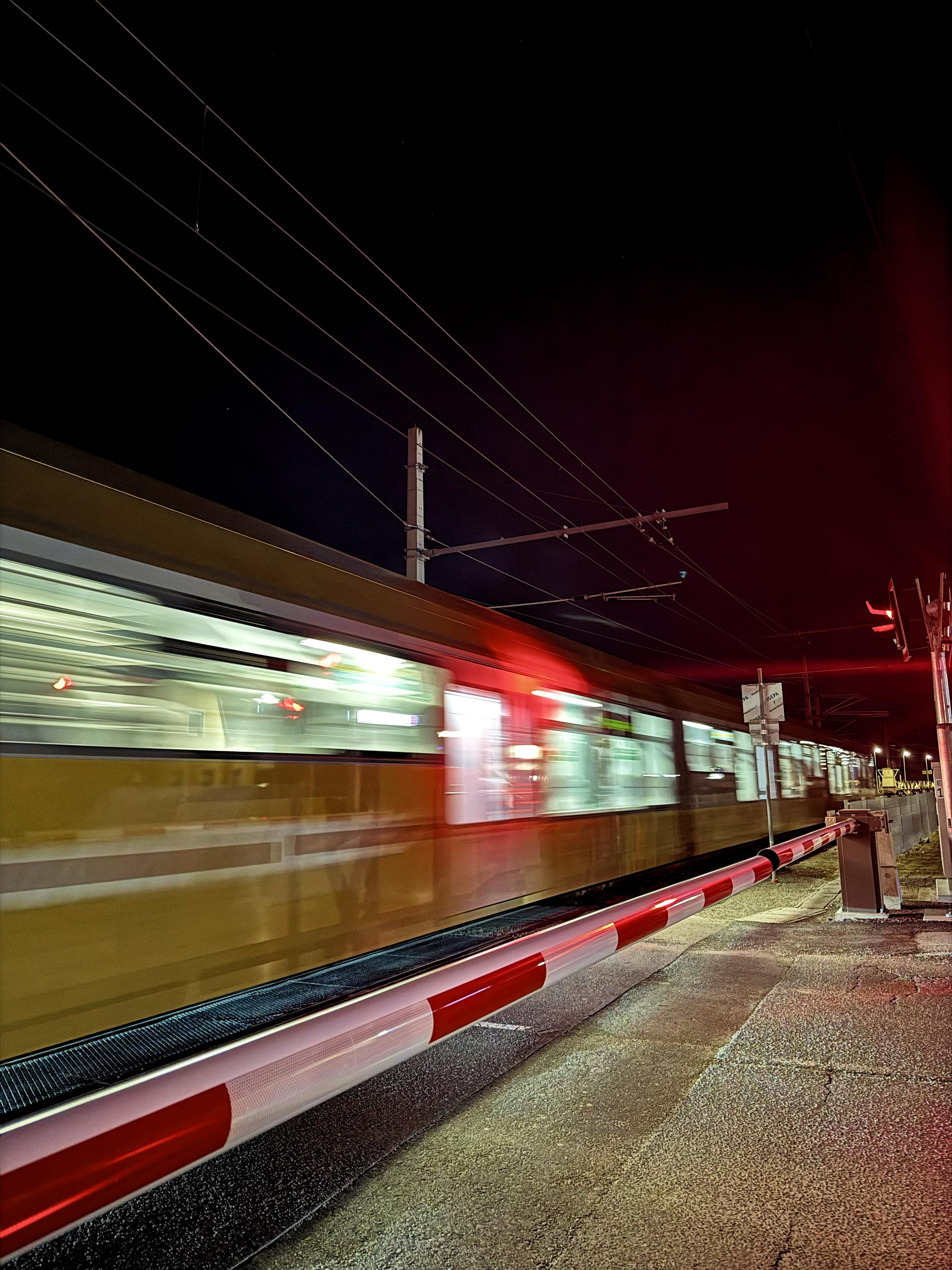 The image shows a train moving quickly through a railway crossing at night. The train is blurred due to its speed, and there’s a red and white barrier in the foreground, indicating the crossing is closed to road traffic. The scene is lit with artificial lighting, likely from both the train and nearby streetlights.