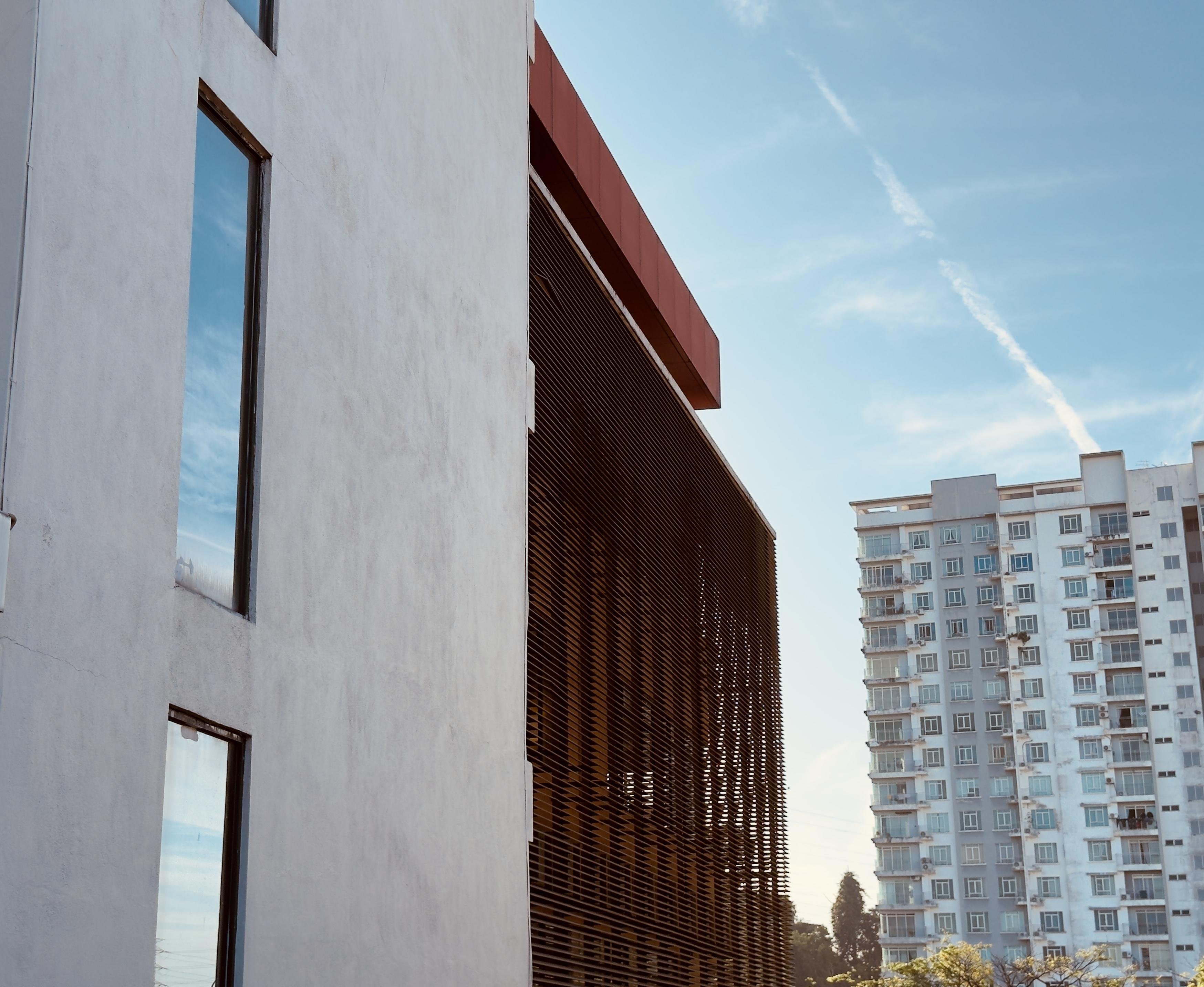 The image shows two buildings. The one on the left is a modern structure with a smooth facade and large vertical windows. The building to the right has a grid-like pattern on its surface. In the background, there is a taller building with multiple floors, featuring a more traditional apartment style with many windows. The sky is clear with some clouds and contrails visible.